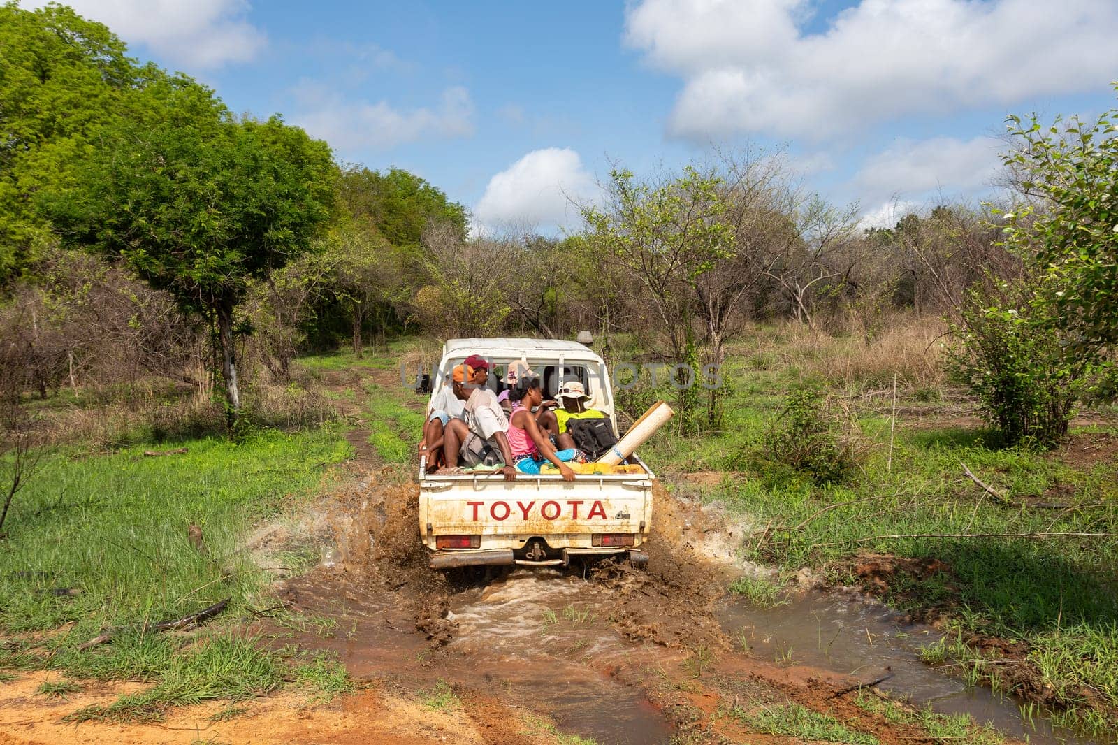 Antsalova, Madagascar - November 7, 2022: Group of people on the back of a car driving through a muddy forest road in a white pickup truck. Standard mode of transport in Africa.