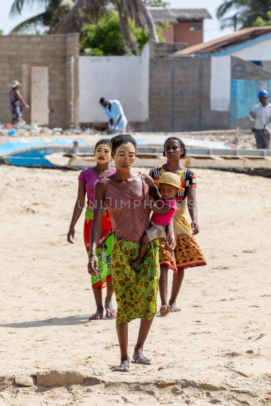 Morondava, Madagascar - November 3. 2022: Malagasy woman with her face painted against the sun with child in her arms walks along the port in Morondava.