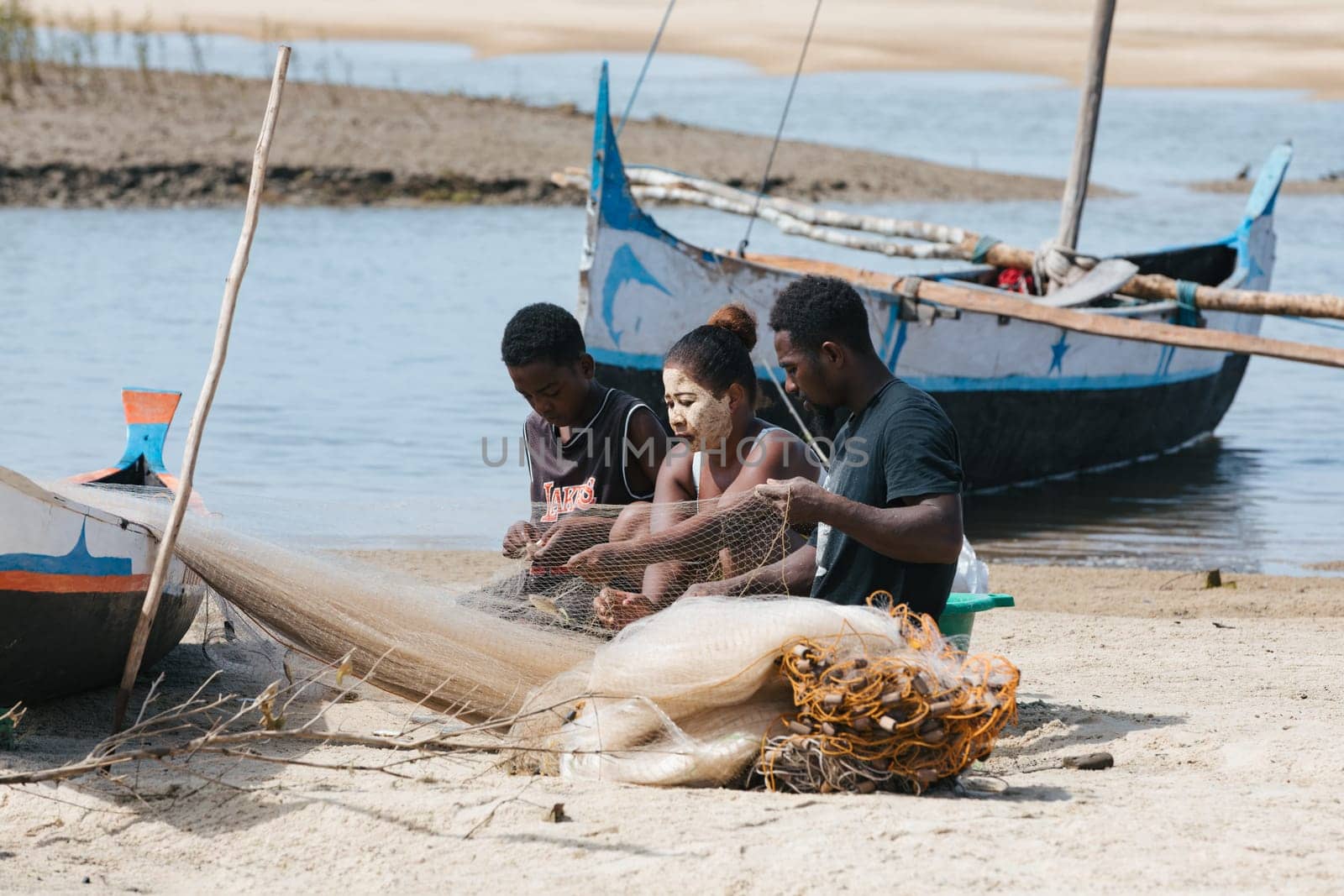 Morondava, Madagascar - November 3, 2022: Fisherman and woman repairing fishing nets at the estuaries of a river, canal Hellot. The woman has a traditionally Malagasy painted face