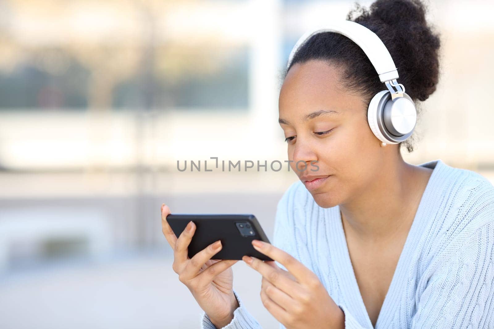 Black woman wearing headphone watching video on horizontal phone in the street