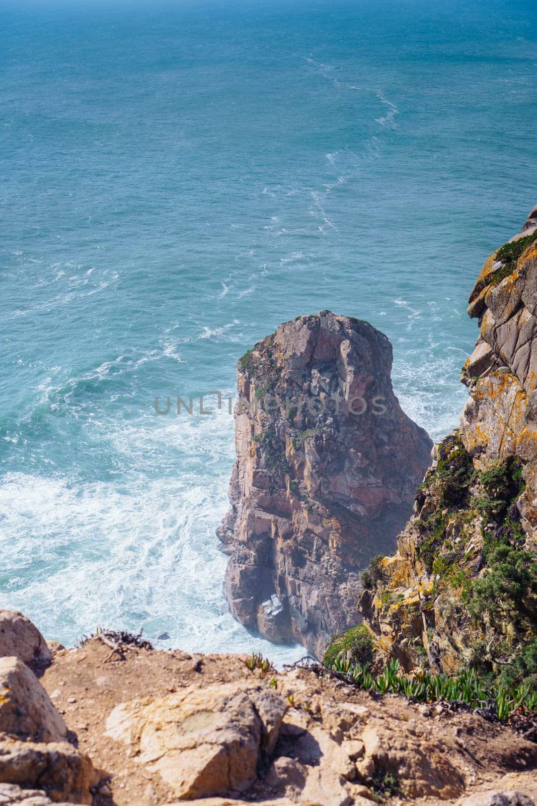 Scenic view of steep rock in blue lagoon near beach seashore coastline surrounded by stormy waves, sheer mountains cliffs covered with green grass. Summer, holiday, travelling, seascape, tourism.