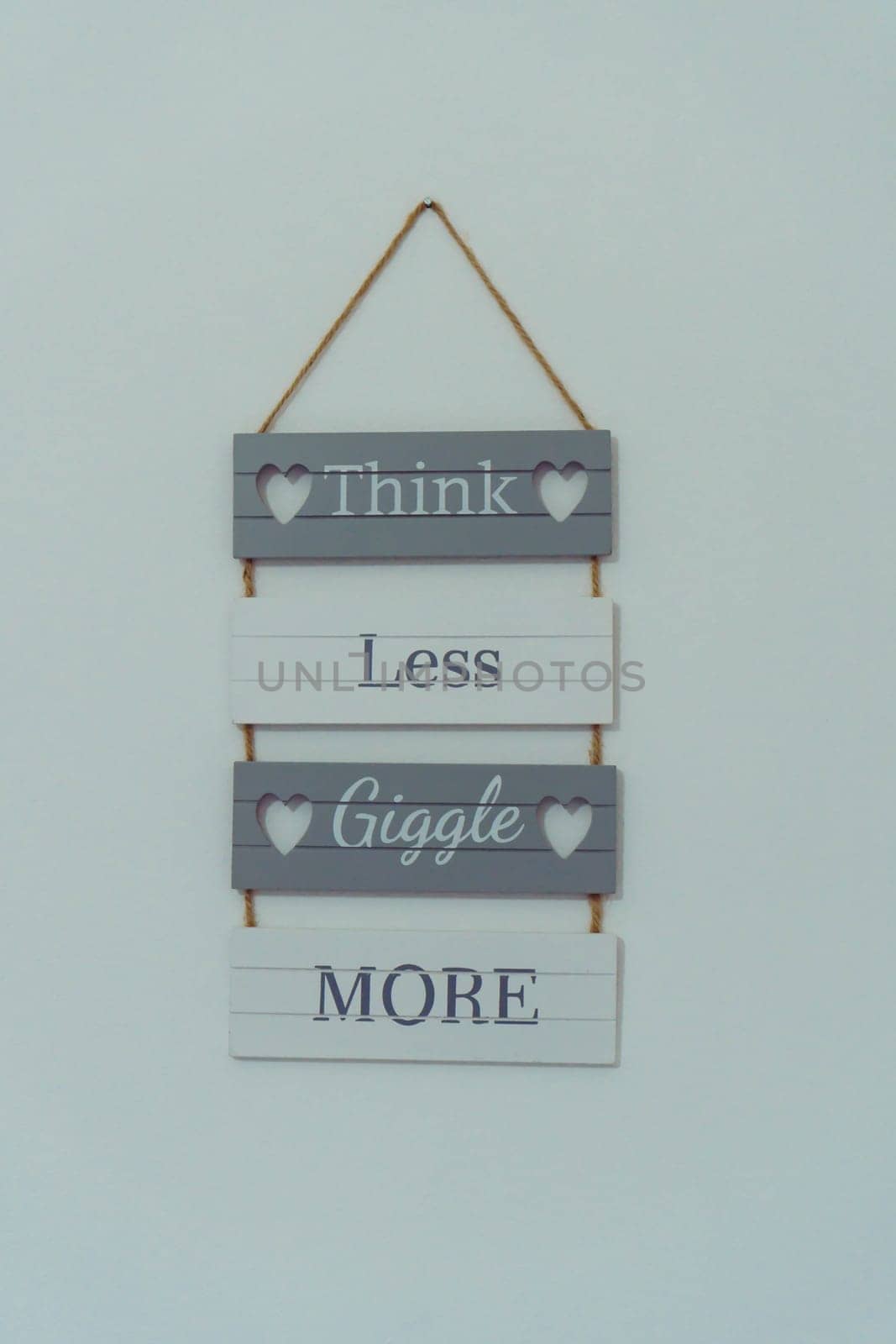 Composition of wooden plaques boards with inscription words think less giggle more, holes in shape of hearts joined with rope hanging on wall on grey background. Wall decoration, placard. Isolated.