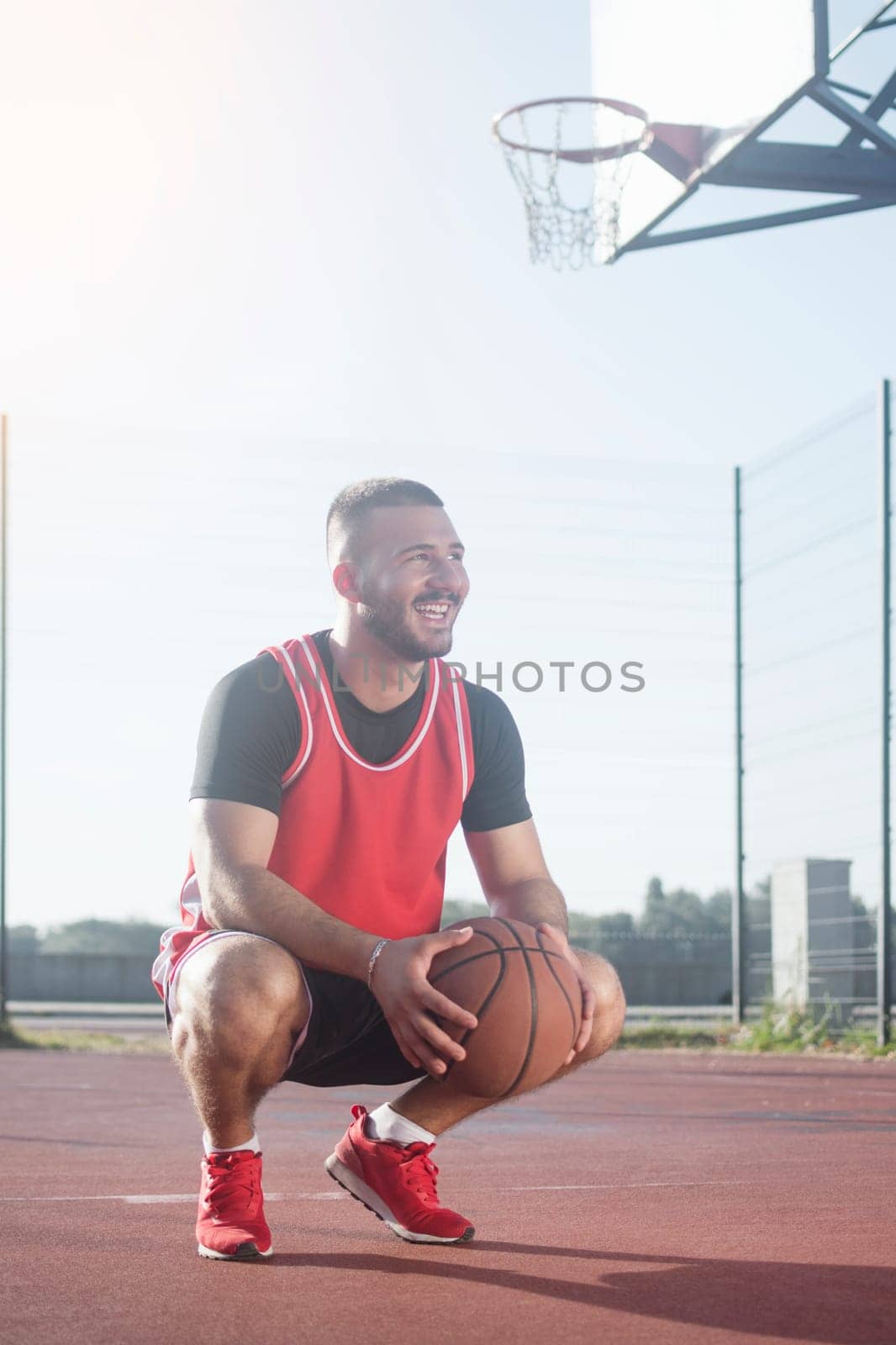 The young smiling basketball player with basketball ball is squatting on playground.