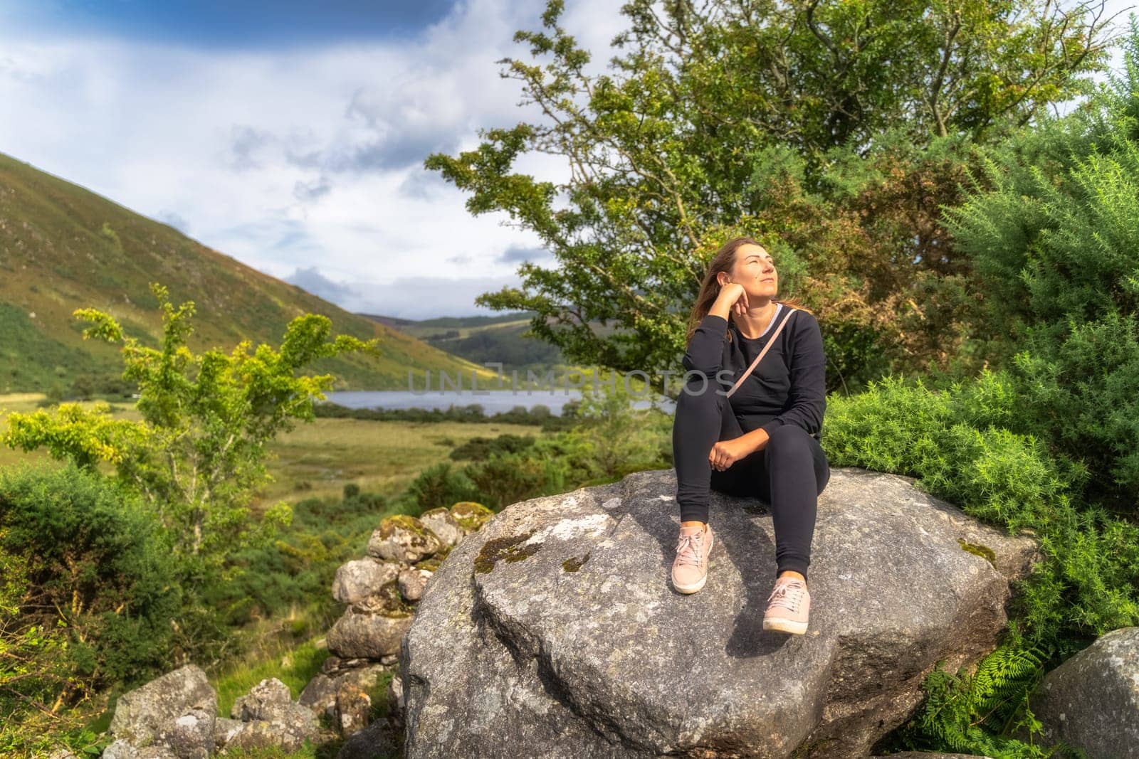 Young woman sitting on a large rock and smiling. Lake Lough Dan and mountain valley in background, Wicklow Mountains, Ireland
