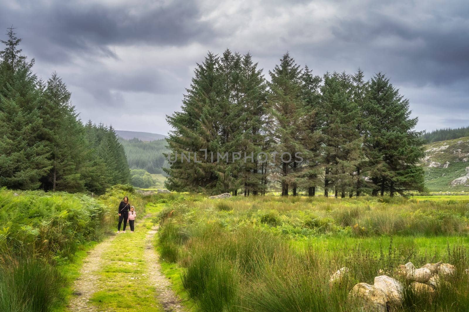 Mother and daughter standing on country road with pine trees and mountain valley in background. Family hike in Lough Dan, Wicklow Mountains, Ireland
