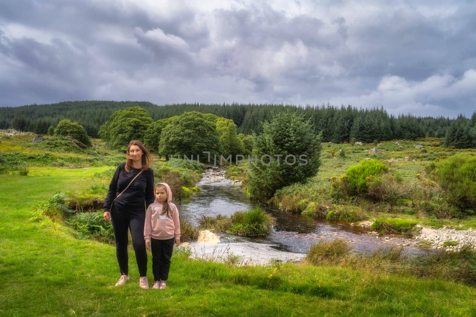 Mother and daughter standing on grass, with winding river and green forest in background. Family hike in Lough Dan, Wicklow Mountains, Ireland
