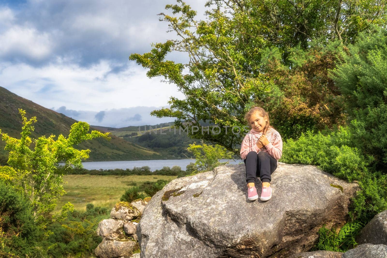 Young girl sitting on a large rock and smiling to camera with lake Lough Dan and mountain valley in background, Wicklow Mountains, Ireland