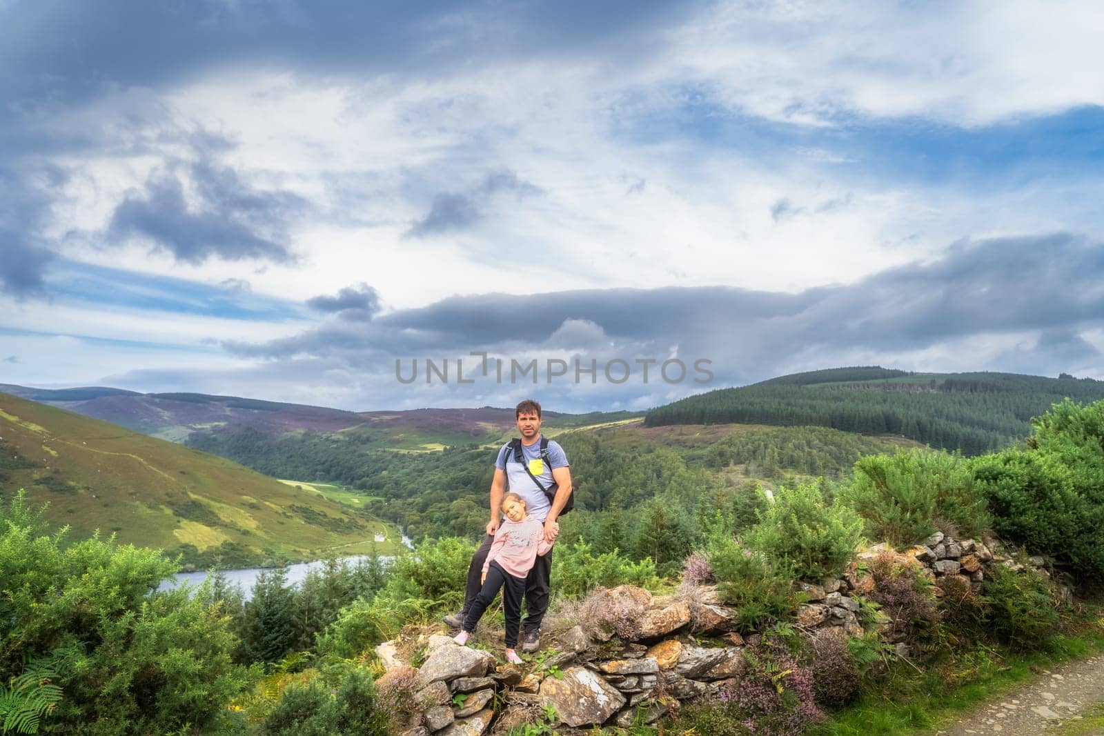 Father and daughter standing and smiling on rock wall. Lake Lough Dan, forest and fields in background. Family hiking in Wicklow Mountains, Ireland