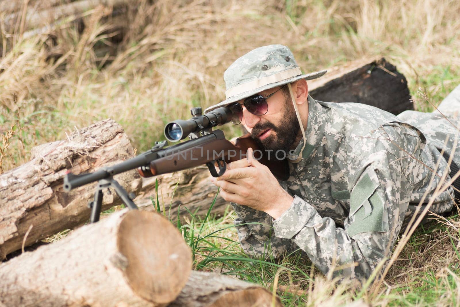 The soldier or hunter in military uniform is lying down, hiding, aiming and shooting a weapon with sniper outdoors.