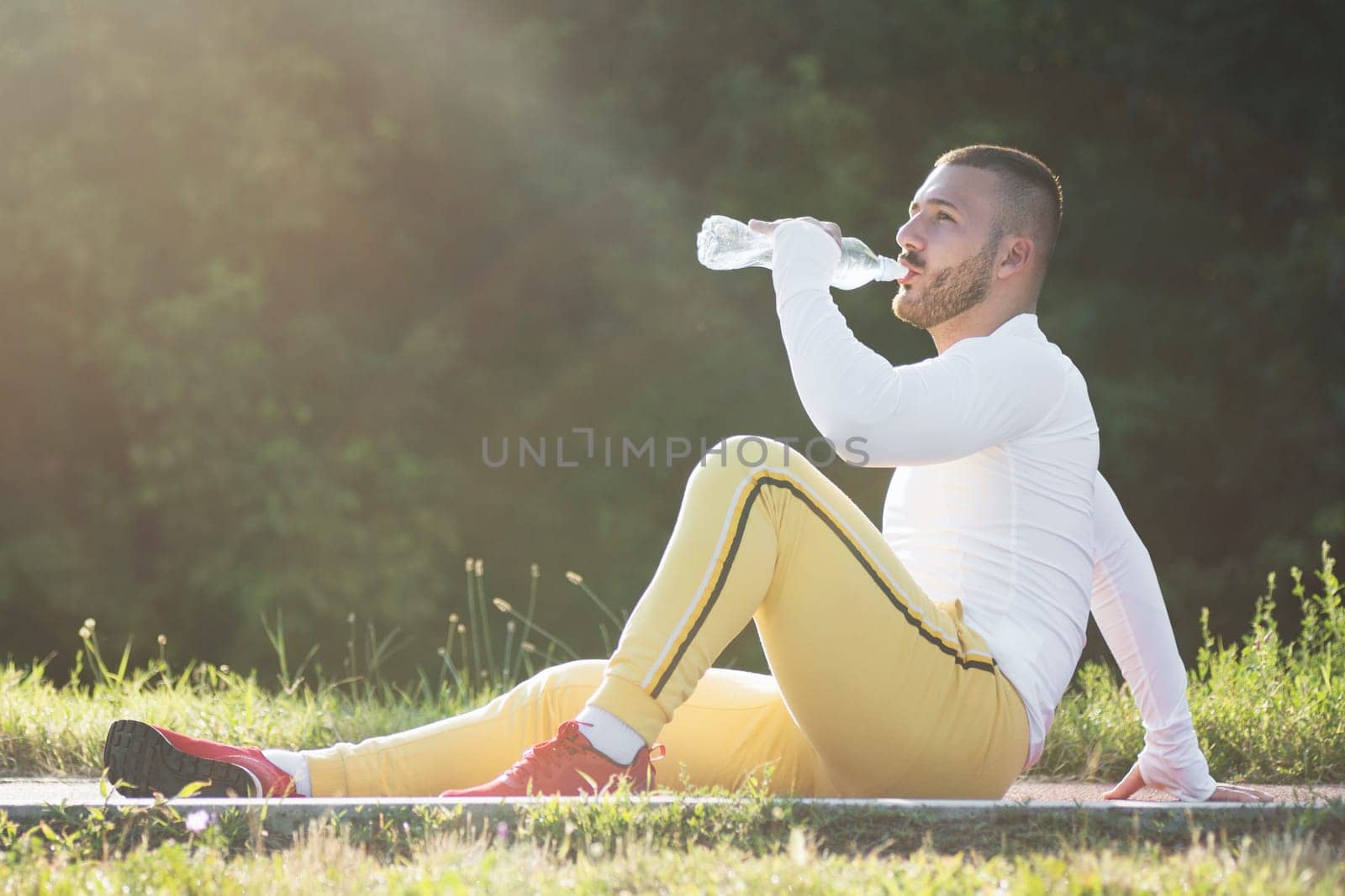 The young sporty guy is drinking water from bottle after jogging or recreation outdoors.