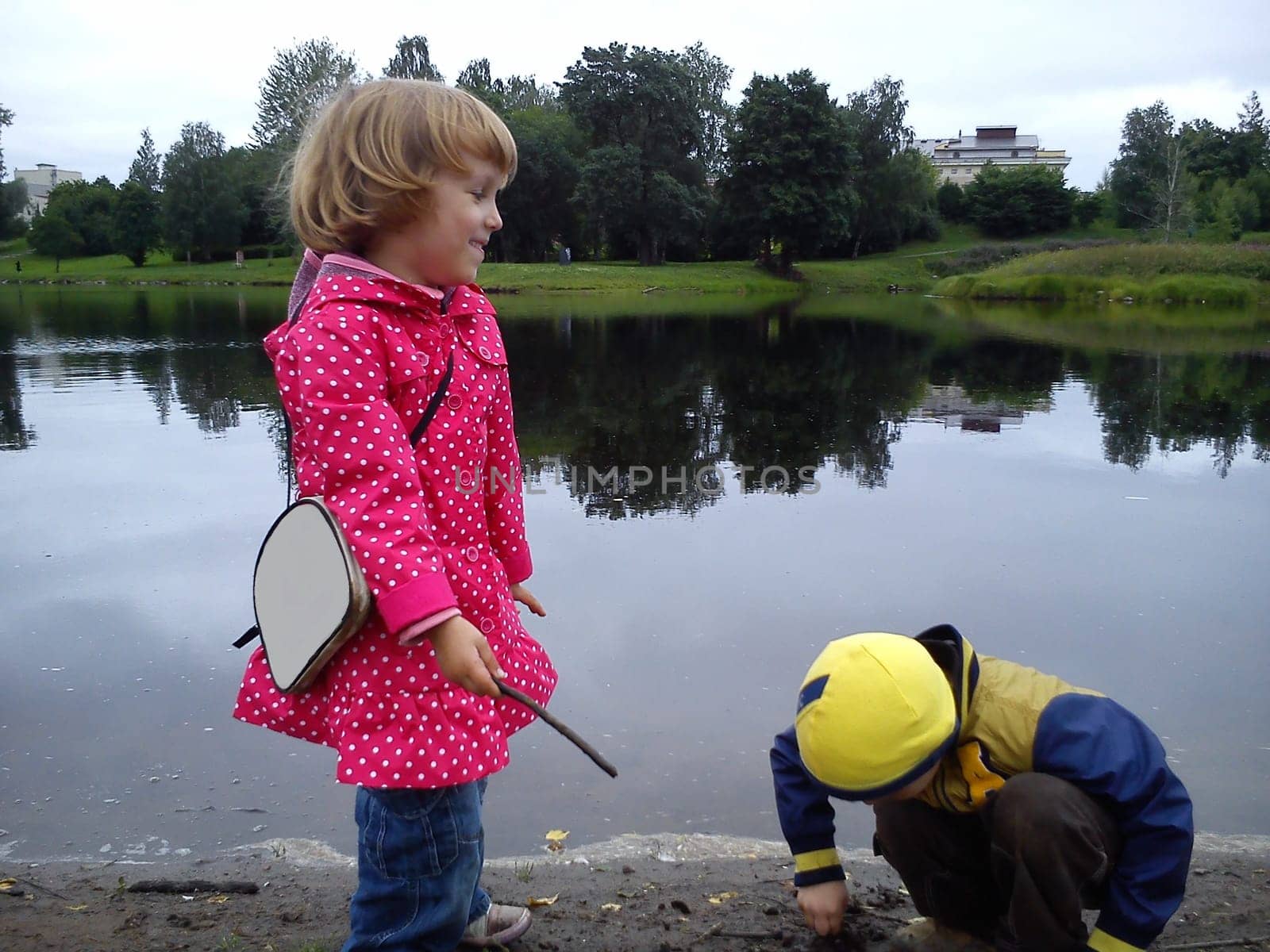 Brother and sister are walking near the river. A boy and a girl are dressed in jeans and autumn clothes. Early summer. Talk and play by the water. Pink cloak in white polka dots, yellow jacket.