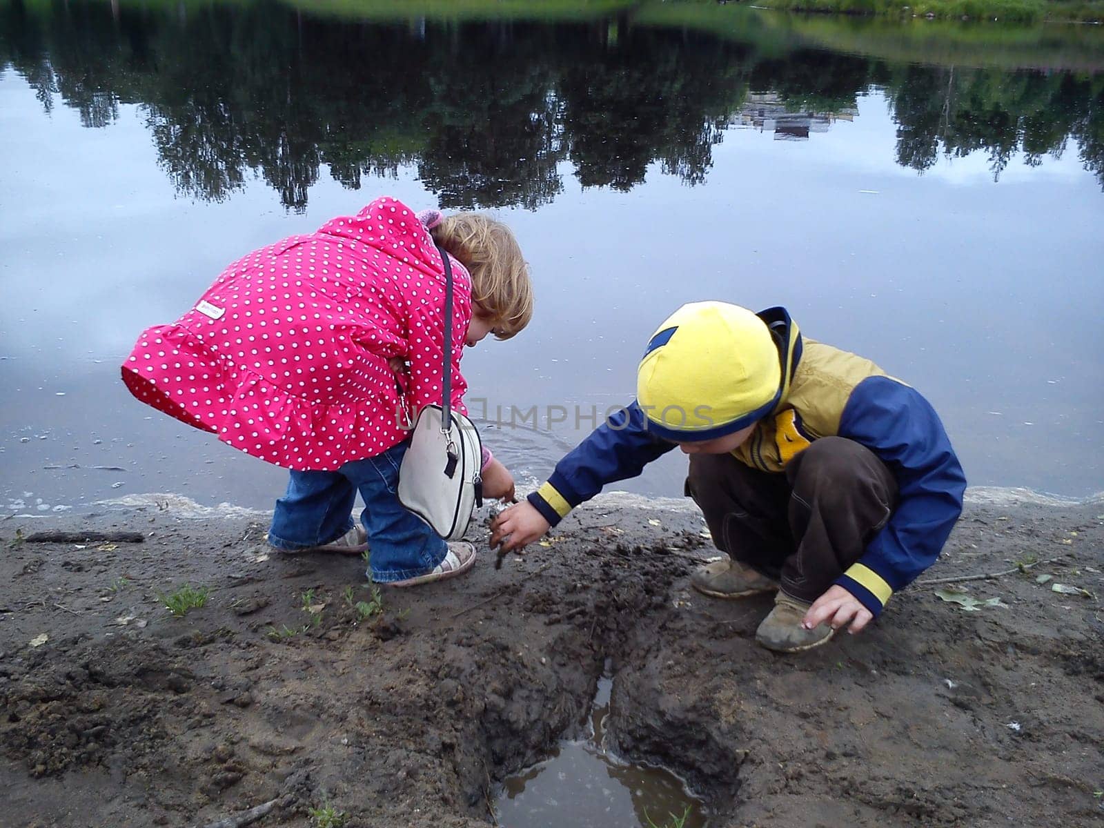 Brother and sister are walking near the river. A boy and a girl are dressed in jeans and autumn clothes. Early summer. Talk and play by the water. Pink cloak in white polka dots, yellow jacket.