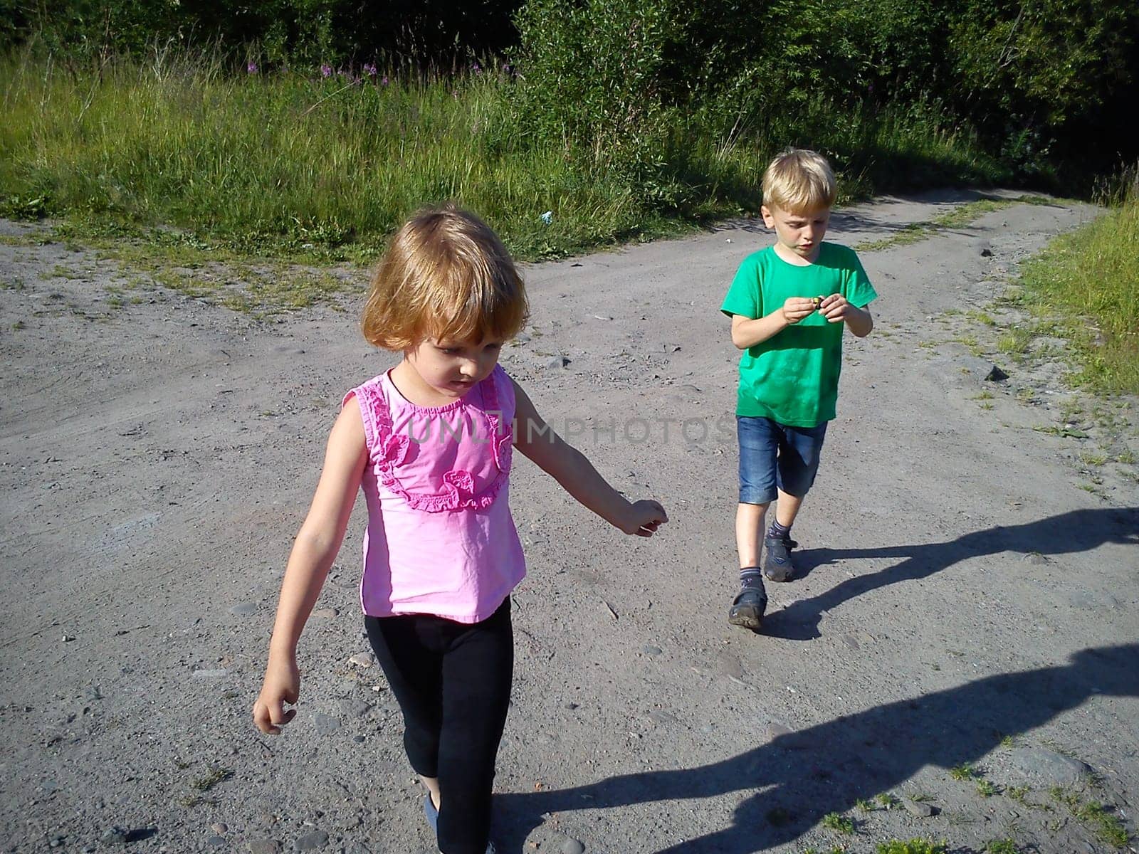 Blond-haired children walk on a dirt road in the summer. Green T-shirt. Girl in a pink blouse with frill. Brother and sister rush home. Hiking in nature in sunny weather. Sweeping movements
