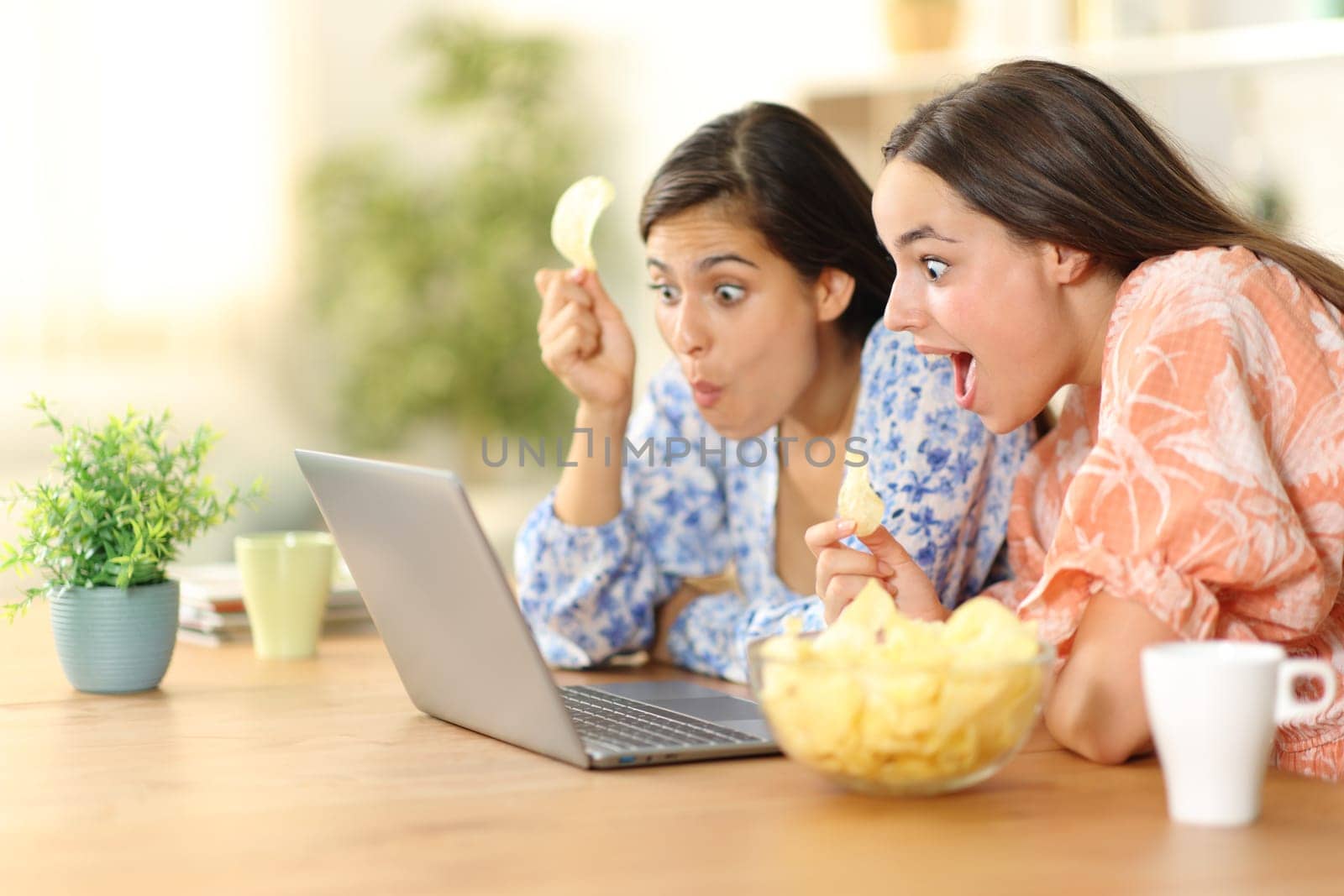 Surprised women watching media on laptop eating potato chips at home