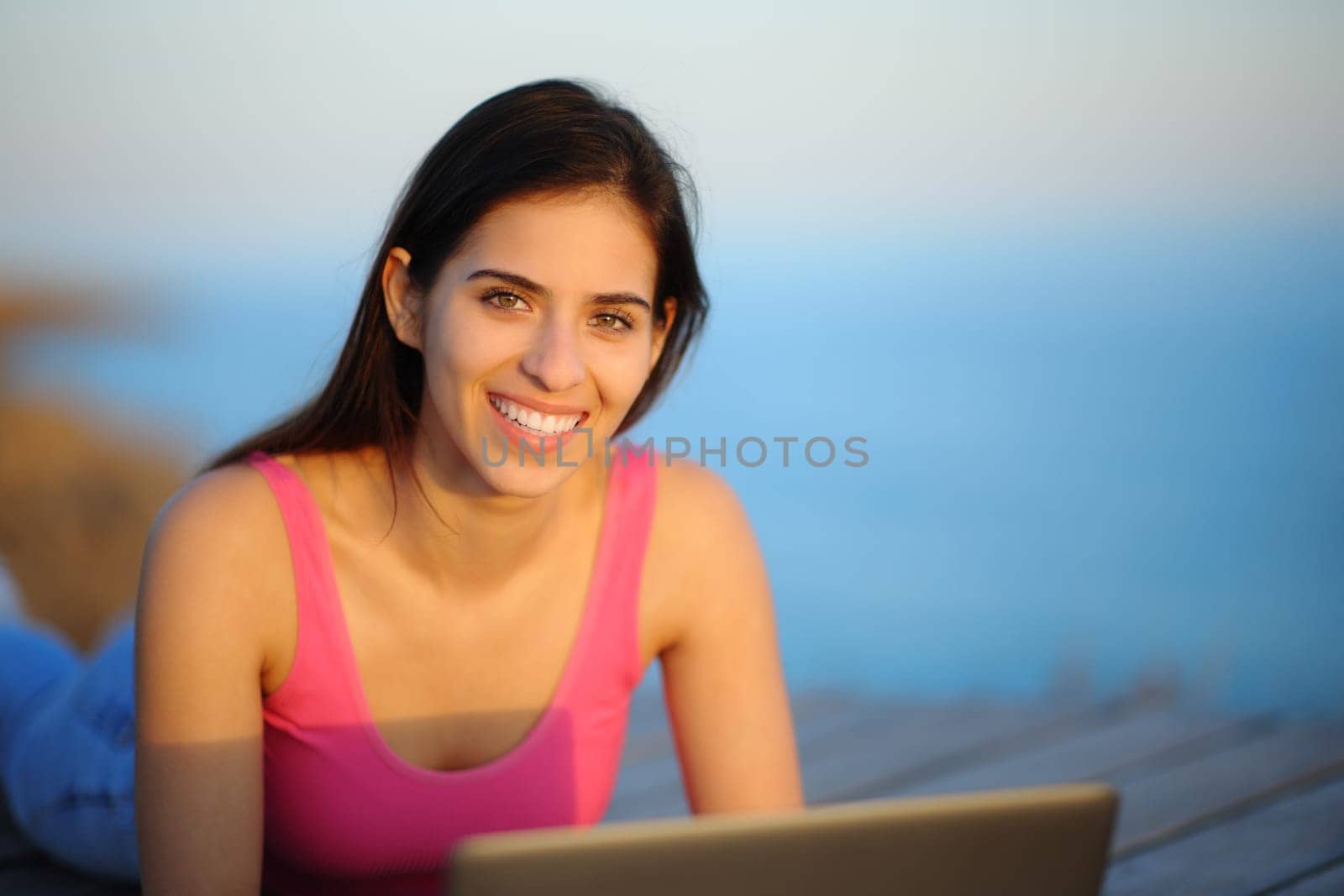 Happy woman in the coast looks at you with laptop at sunset