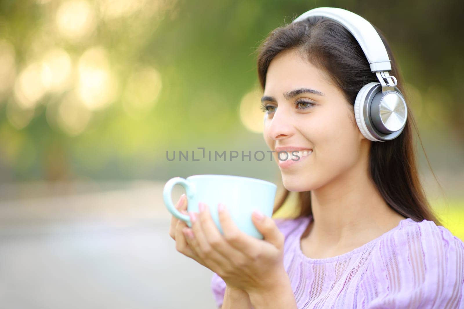 Happy woman listening to music drinking coffee looking away in a park