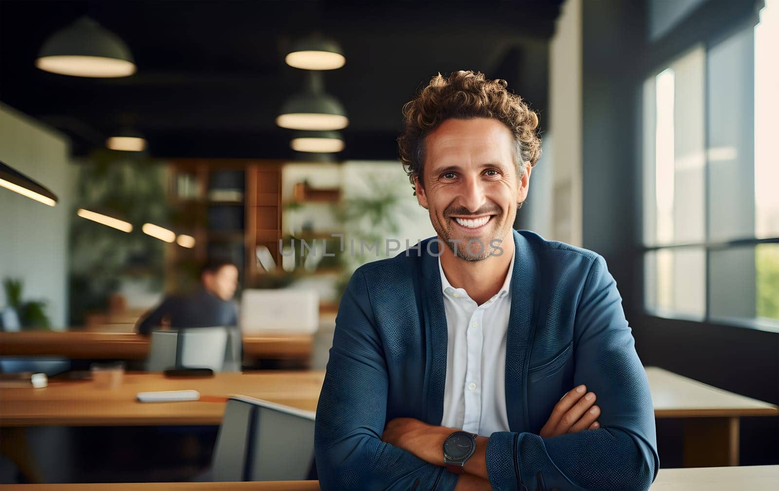 Portrait of young beautiful man smiling with crossed arms, looking at camera. Joyful, cheerful, happy employee sitting at desk or workplace in modern office. Successful businessman or manager