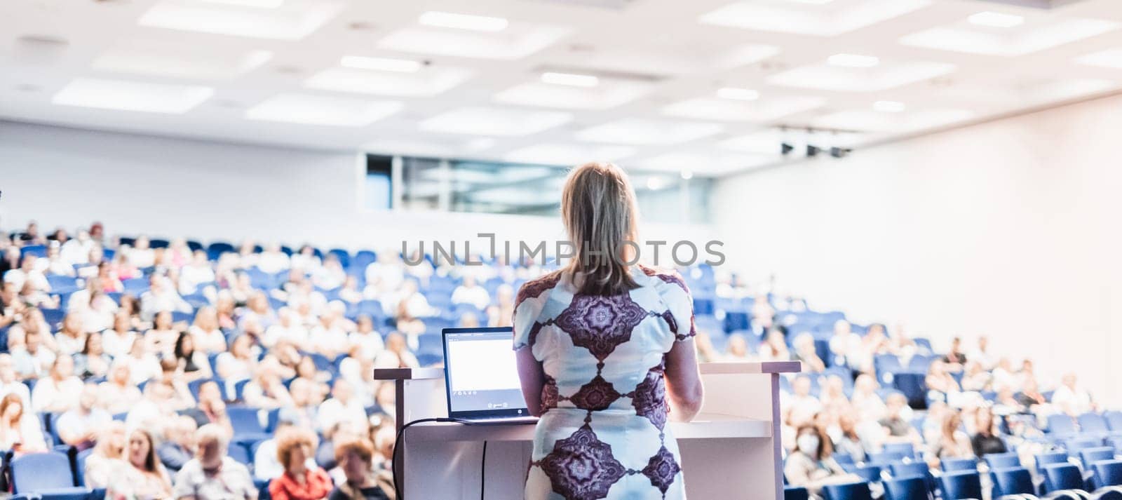 Female speaker giving a talk on corporate business conference. Unrecognizable people in audience at conference hall. Business and Entrepreneurship event.