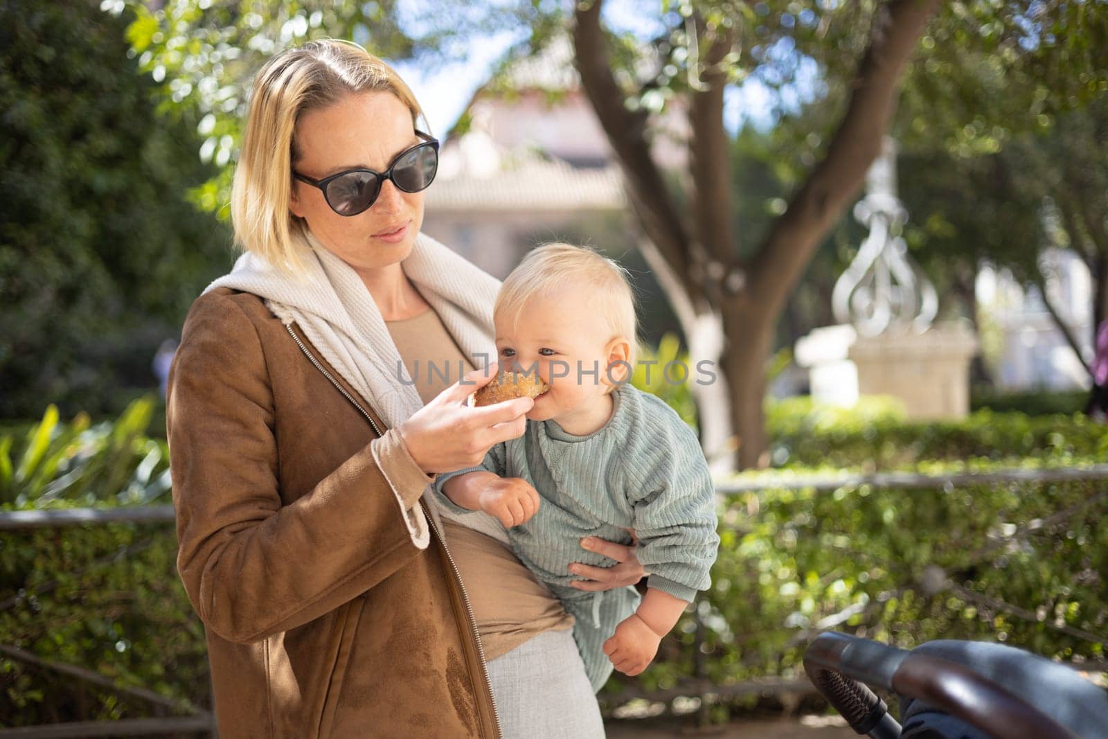 Close up portrait of sweet toddler kid eating fruit puree from plastic doy pack, sitting in stroller, outdoor snack time