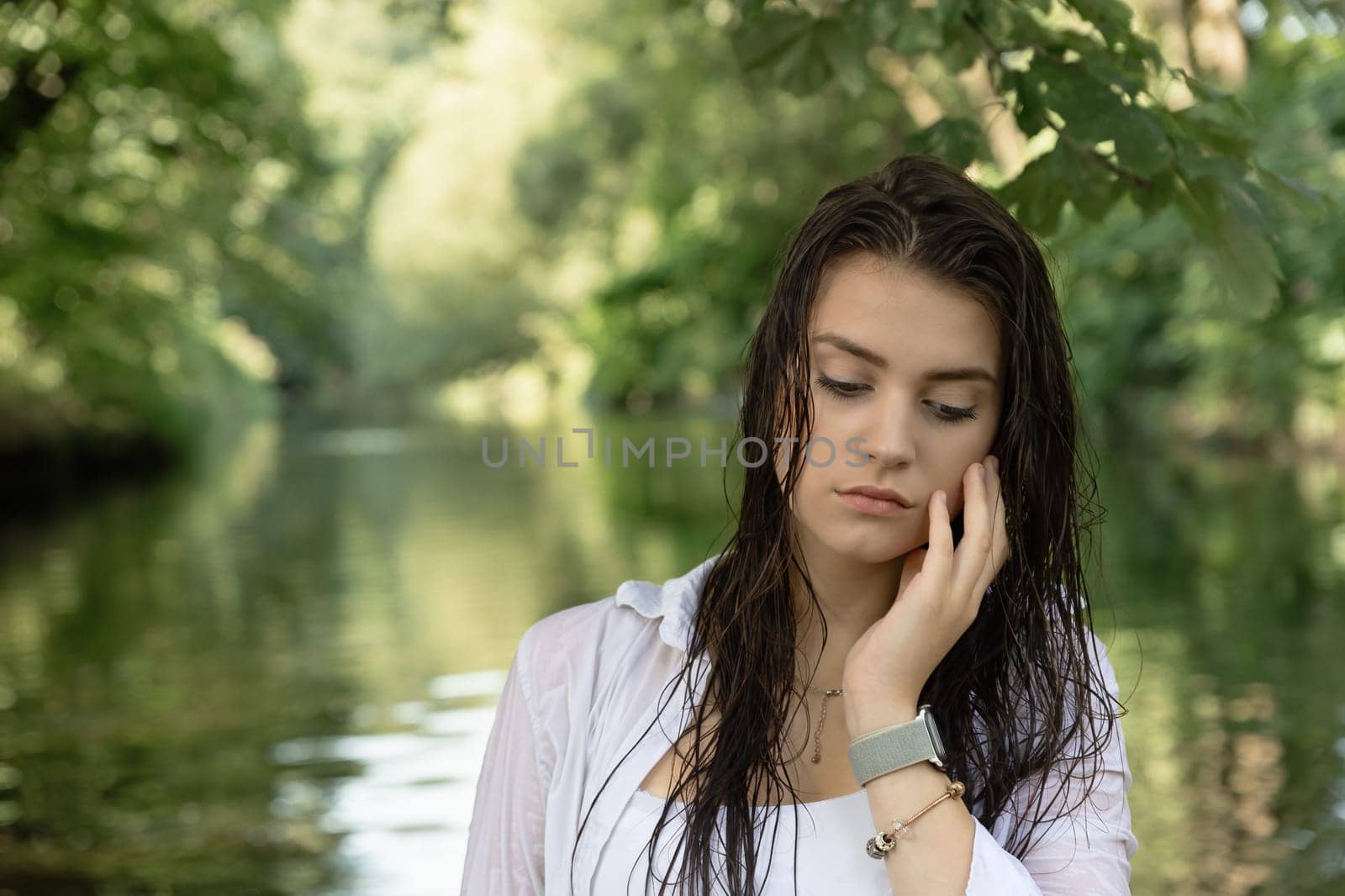 Sad brunette girl with a hand supporting her face is posing with wet hair in the river. Horizontally.