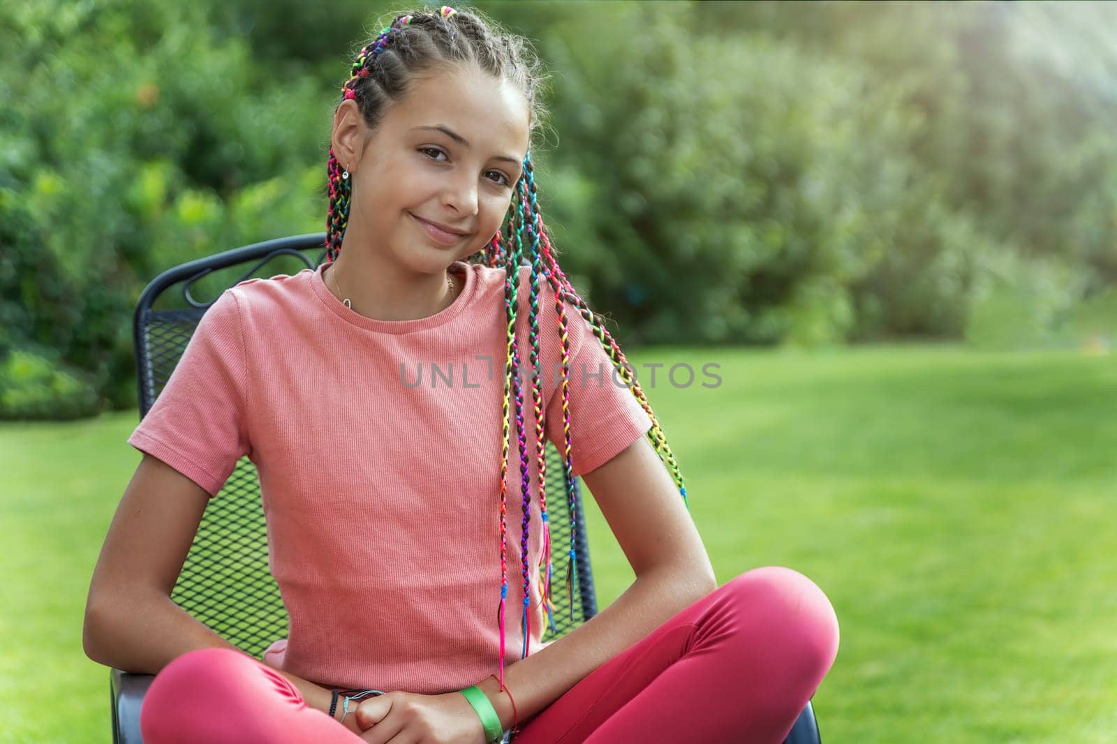 Portrait of smiling young girl with colorful braids in her hair sitting on a chair outside. Horizontally.