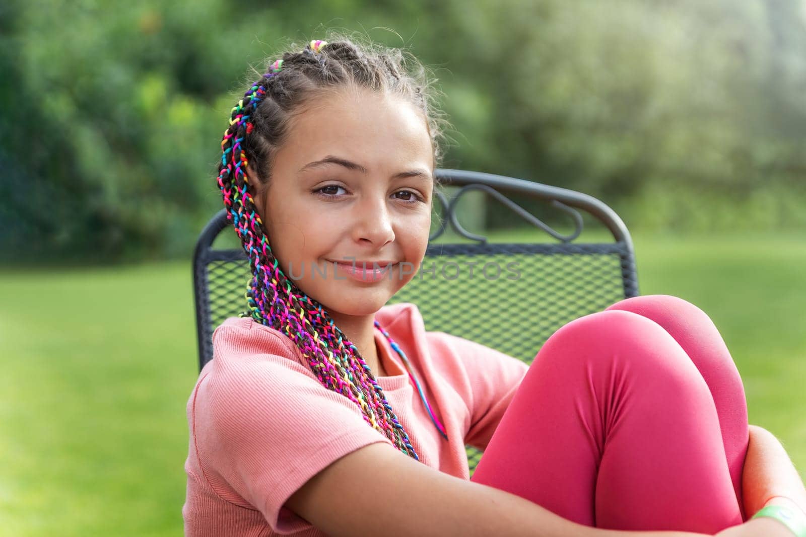 Side view of young girl with colorful braids in her hair sitting on the chair outside. Horizontally.