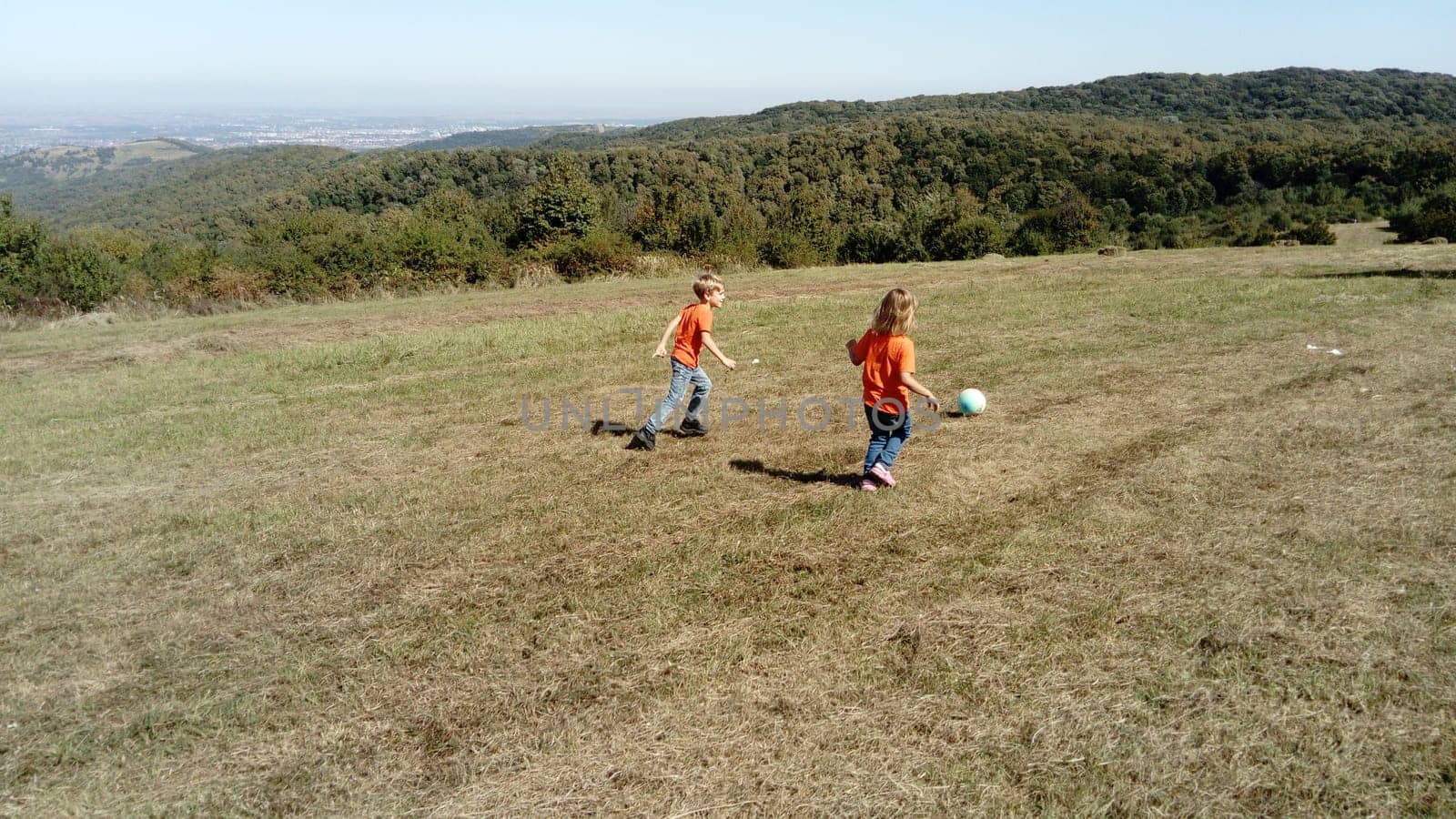 A boy and a girl with blond hair, dressed in orange t-shirts and classic jeans, play ball in a mountain meadow. Early warm autumn. In the background, the horizon and vegetation