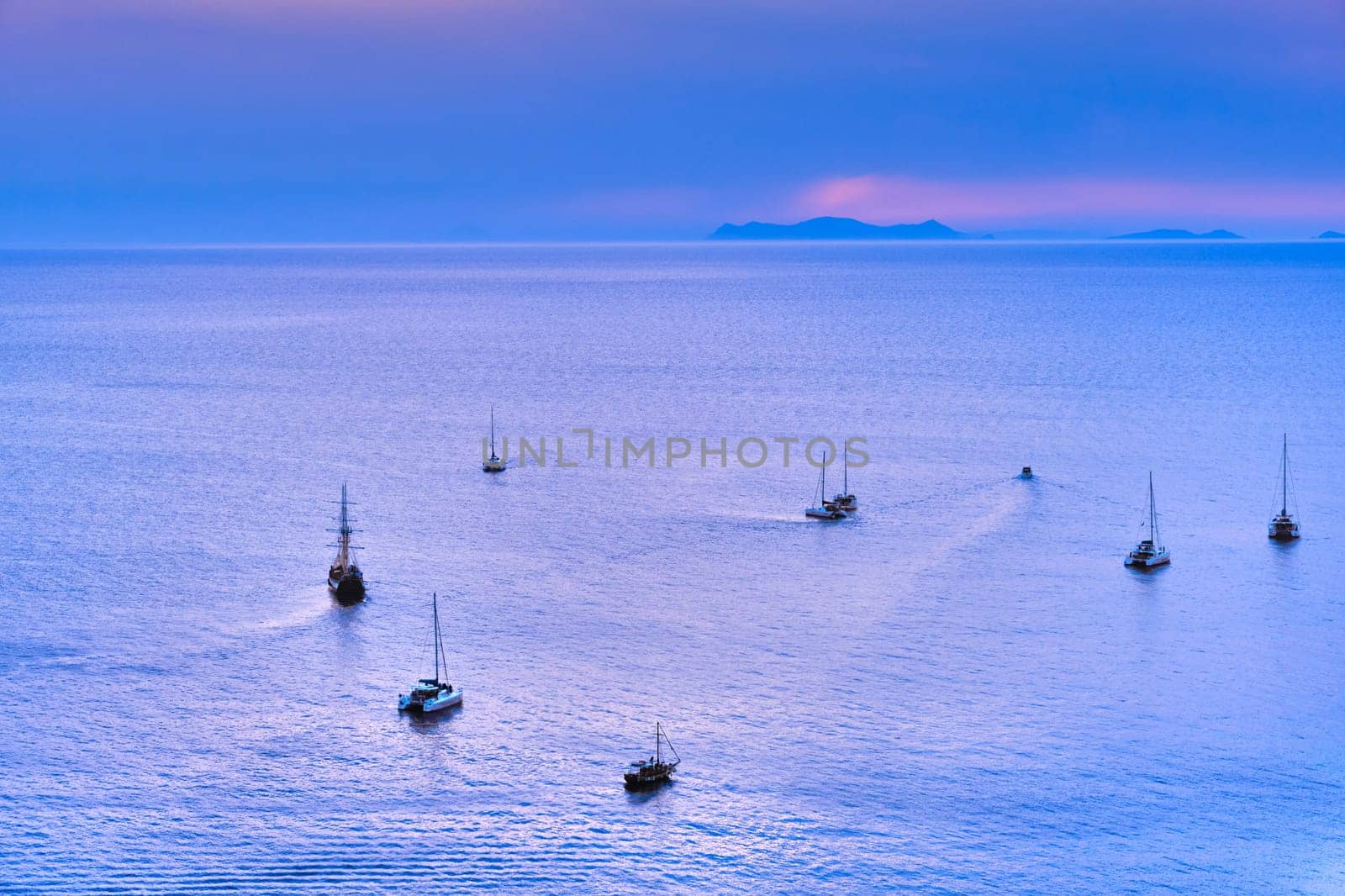 Aerial view of numerous sailboat and yachts go to sea for sunset or sunrise. Calm sea waters, hazy distant sun rays, blue sky and clouds, seascape, cloudscape. Adventure, summer vacations, tourist attraction.