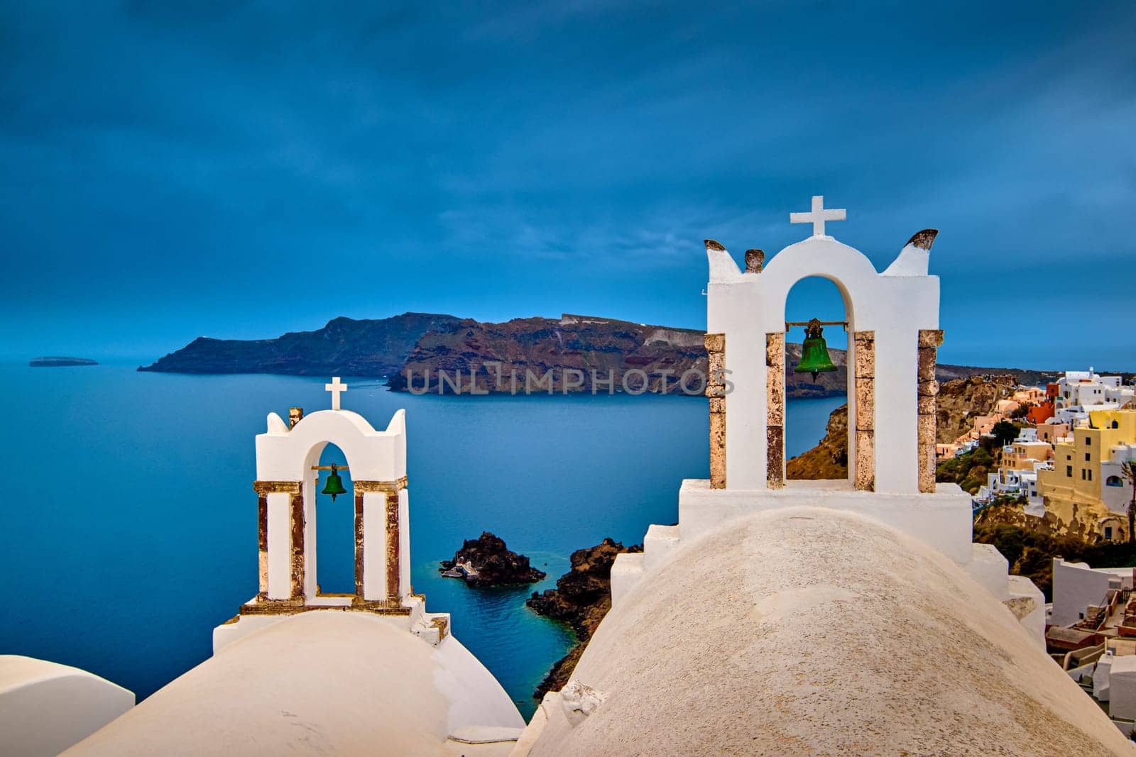 Two bell towers and dome roofs of white Greek Orthodox church in Oia village, Santorini island, against waters of Aegean sea, Greece, and distant island on overcast day with heavy clouds and dramatic sky. Famous local landmark, wedding destination