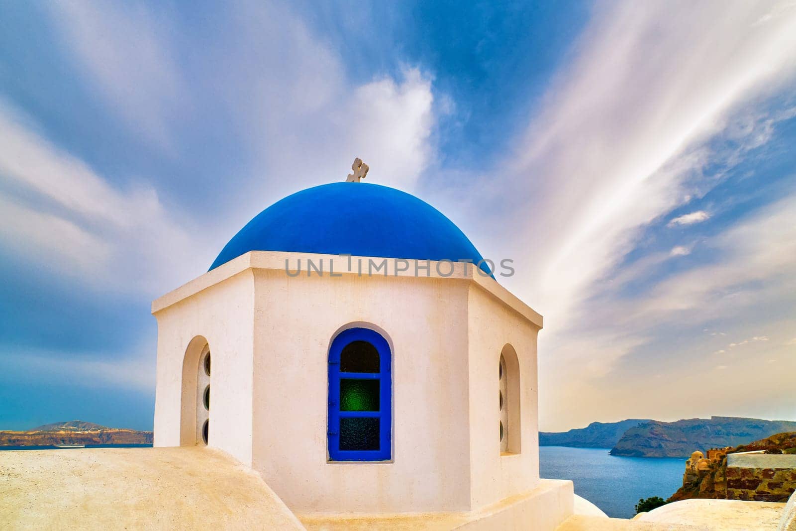 White Greek Orthodox church and its blue dome with cross against dramatic blue sky and clouds, Santorini island, Greece. Beautiful landscape, sea waters, islands, harbor, cliffs, travel destinations in Mediterranean sea cruises, tourist attractions