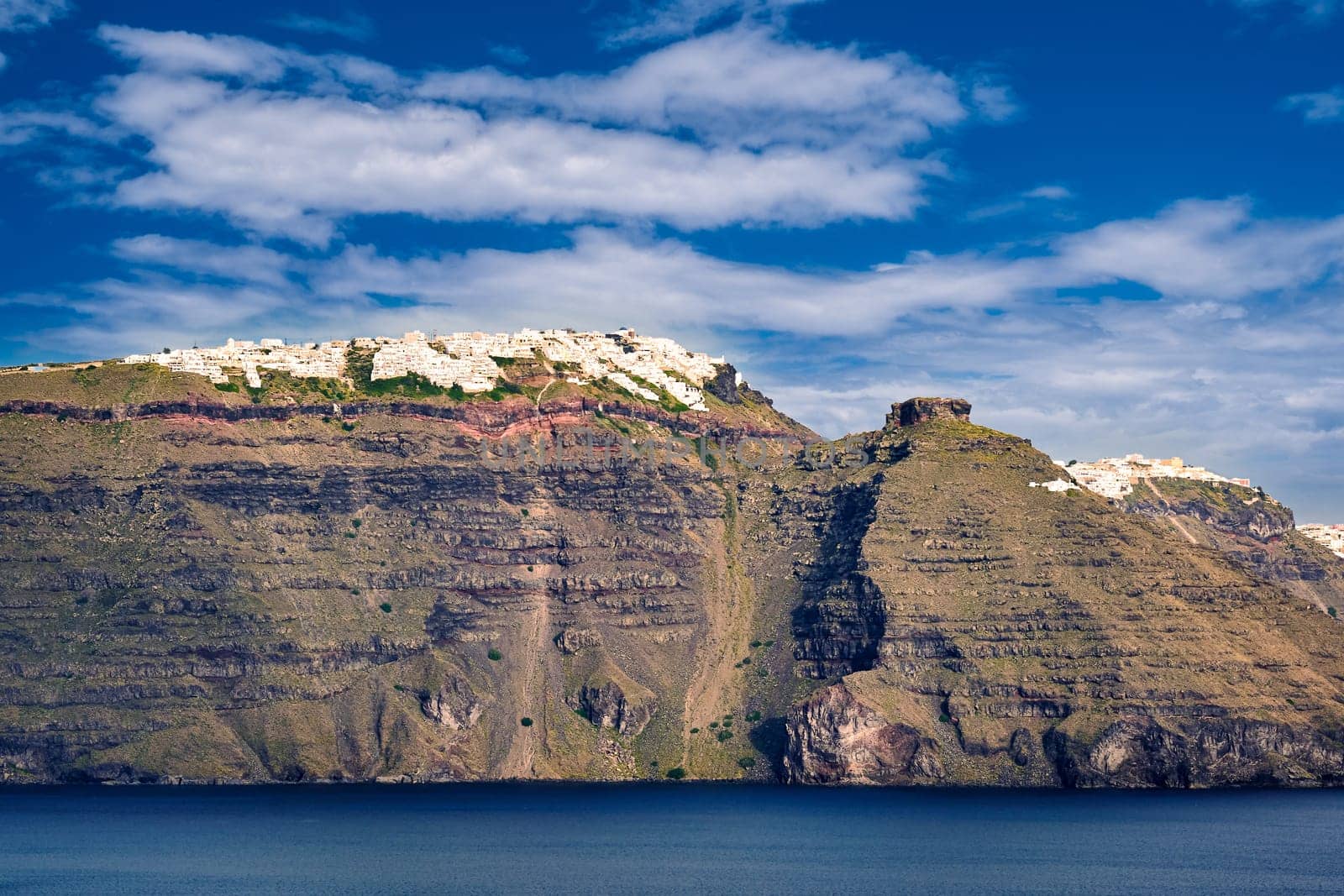 Panorama of Santorini island, Greece. Rough cliffs of volcanic island, sunny day, caldera bay, blue sky and clouds, whitewashed town on hilltop, summer Mediterranean cruise, vacations, travel destination, tourist attraction, clear Aegean sea waters