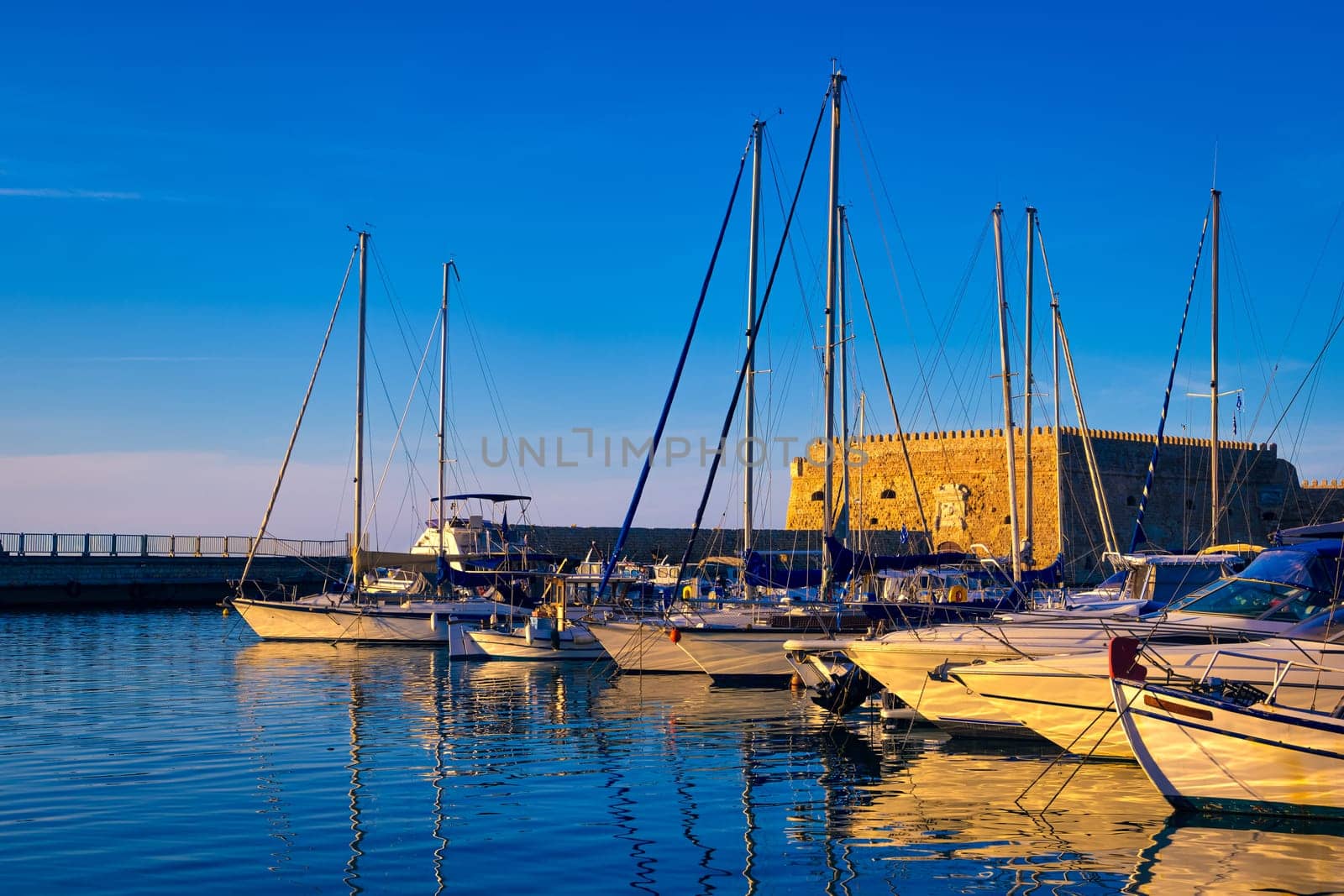 Landscape of Venetian castle or Koules fort in Heraklion, capital of Crete island, Greece and moored fishing boats at sunset. Clear blue sky, deep blue waters of port, yachts, sailboats. Travel destination of Mediterranean sea, summer vacations.