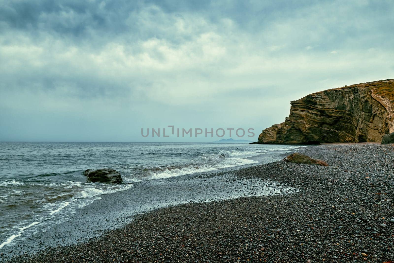 Dramatic landscape of stormy sea or ocean waves rolling on rocky volcanic beach on cloudy day. Overcast sky, heavy grey clouds, black lava pebbles and sand, weathered cliff. Santorini island, Greece.