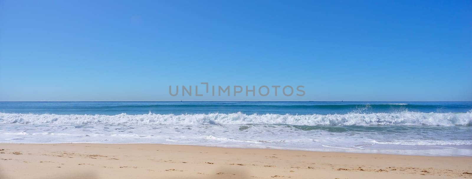 Blue sky, breaking surf waves and sandy beach on a calm sunny day on the water's edge as the tide comes in along the shoreline of Kawana Beach, Sunshine Coast, Queensland, Australia