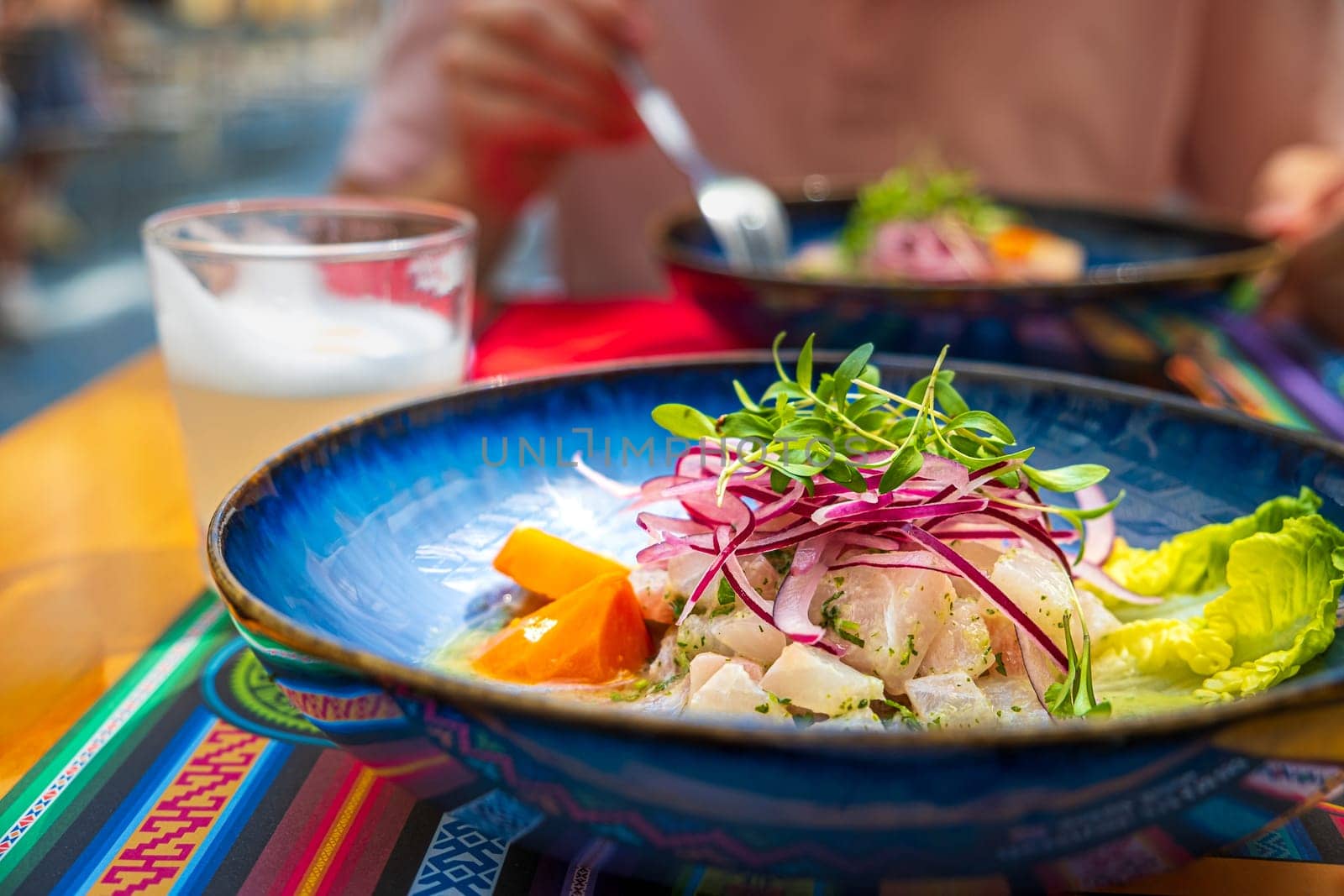 Fresh ceviche served in blue plate over placemat with traditional patterns, Pisco Sour cocktail. Unrecognizable adult in blurred background. Peruvian-style ceviche made out of raw fish, red onions, corn, roasted corn and sweet potato. Selective focus