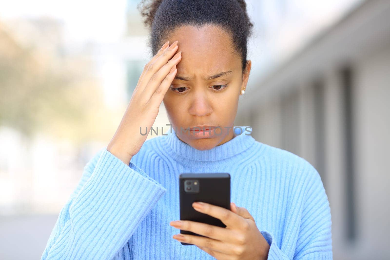 Front view portrait of a worried black woman checking bad news on phone in the street