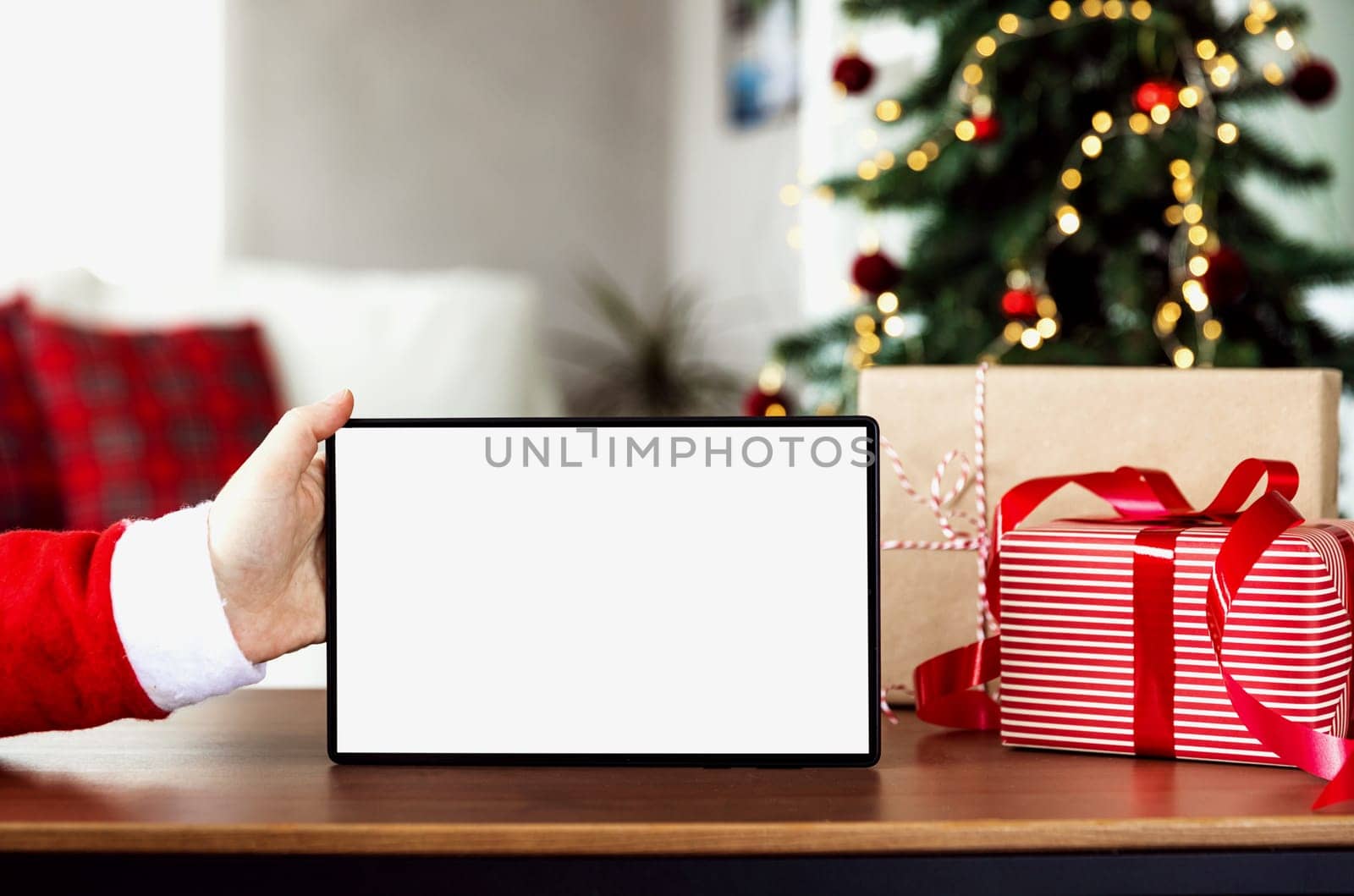 Christmas, New Year background with copy space for design. Santa hand holding empty blank white tablet screen. Mockup for product placement montage. Online shopping from home. Fir tree on background