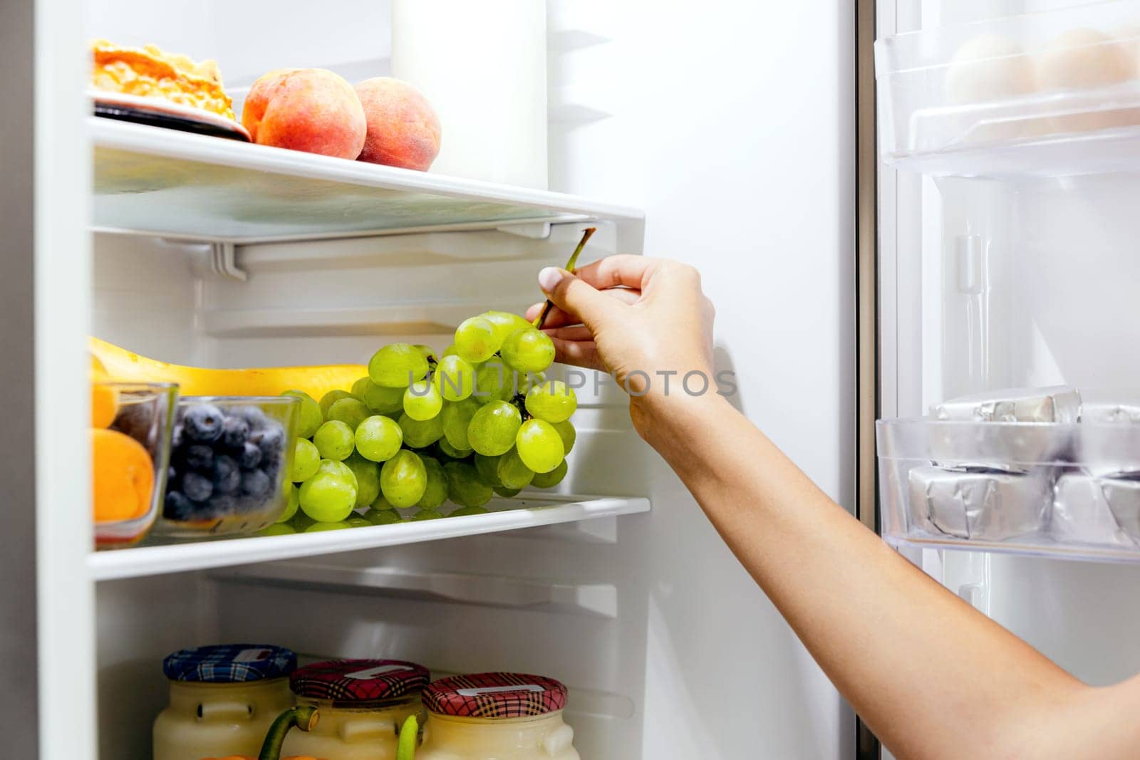Woman hand taking, grabbing or picks up green bunch of grapes out of open refrigerator shelf or fridge drawer full of fruits, blueberries, bottles with yogurt. Healthy food diet, lifestyle concept