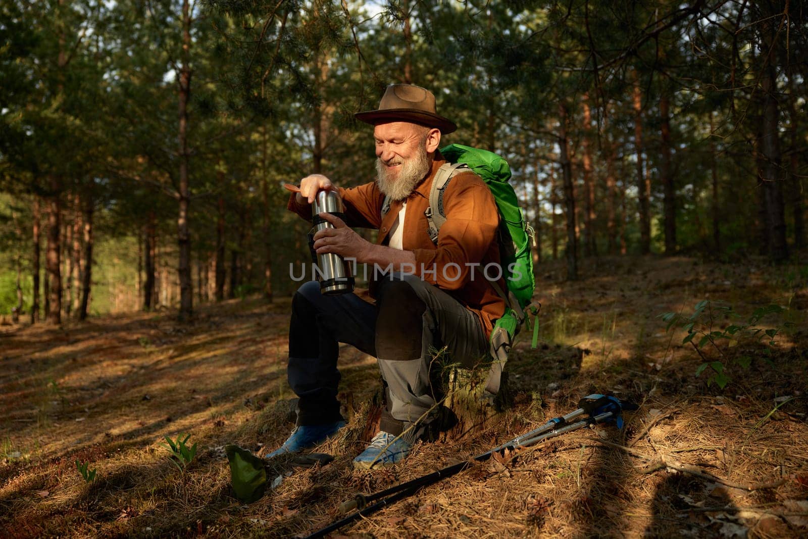 Senior man hiker drinking warm tea from thermos cup during rest in forest. Hiking outside on retirement