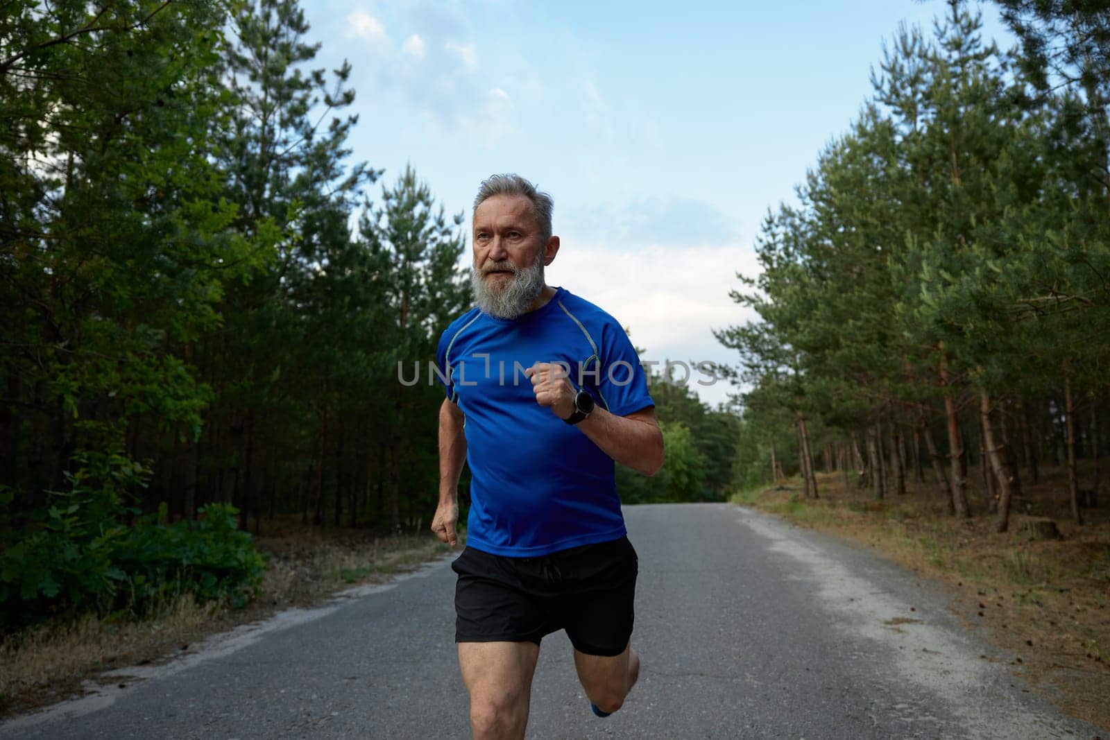 Senior sportsman running among countryside road. Physical endurance training and cardio exercise for pensioner