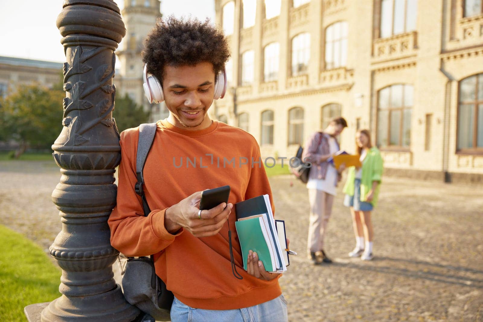 Carefree relaxed teenager boy student in earphones using mobile phone to listen music standing outdoors. Break time at high school, university or college