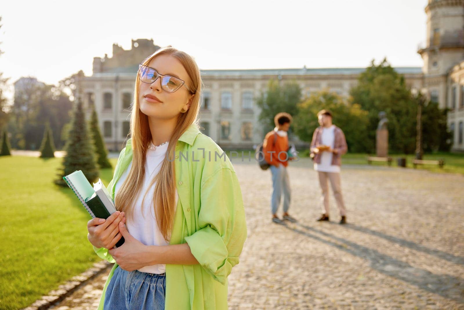 Outdoors portrait of smart female student wearing eyeglasses and casual clothes holding studying accessories standing over university campus yard with classmates on background