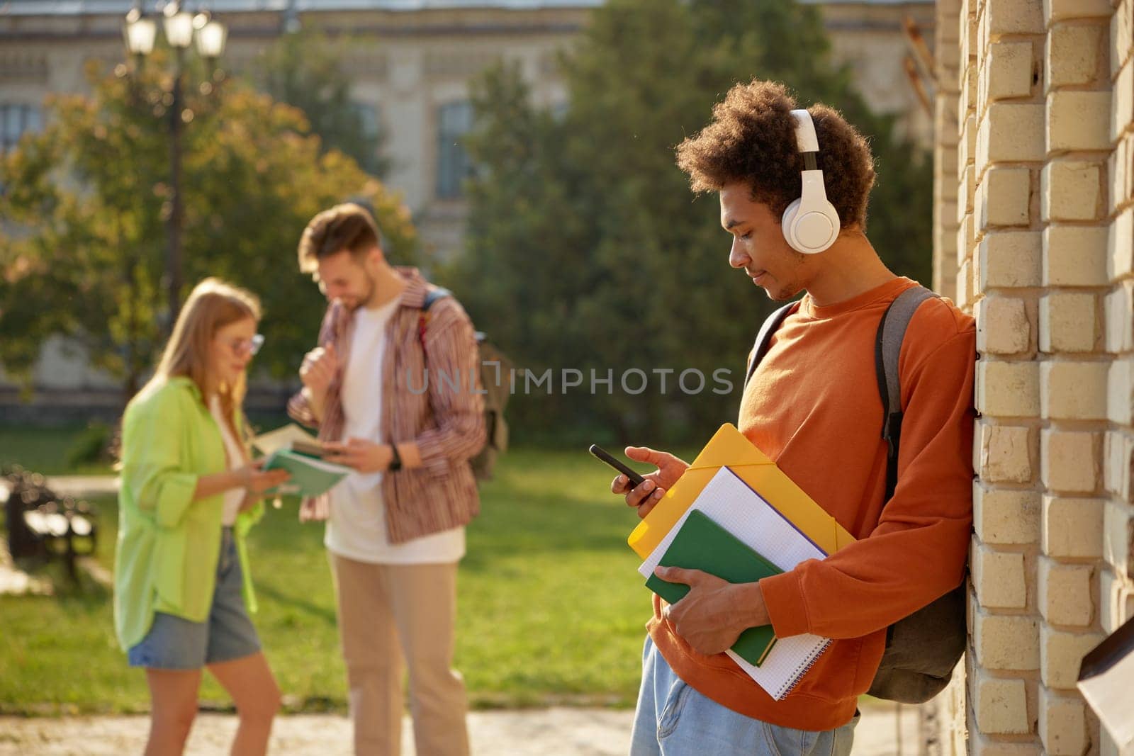 Teenager students at university campus after day of class. Young guy in headphones using mobile phone standing with textbooks in foreground and classmates friendly talking on blurred backdrop