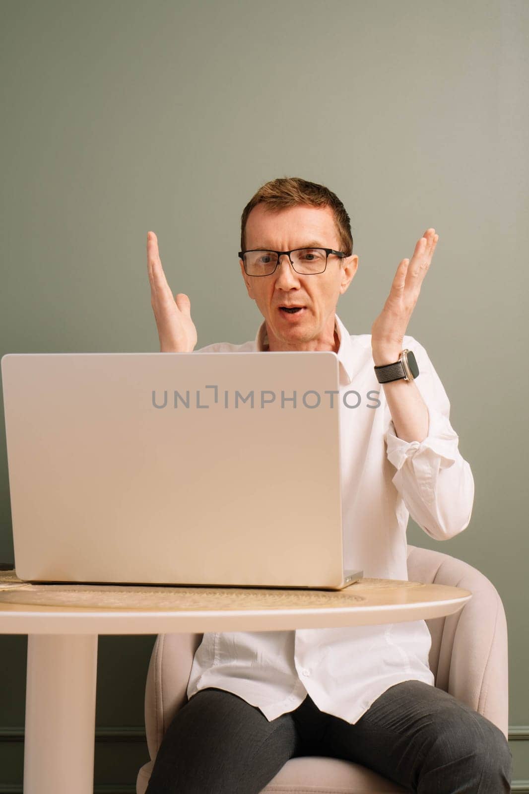 Stunned middle aged man furiously raised hands while working on laptop from home office. Workman in glasses dissatisfied with work, bemused by result, look at computer monitor while sitting at table.