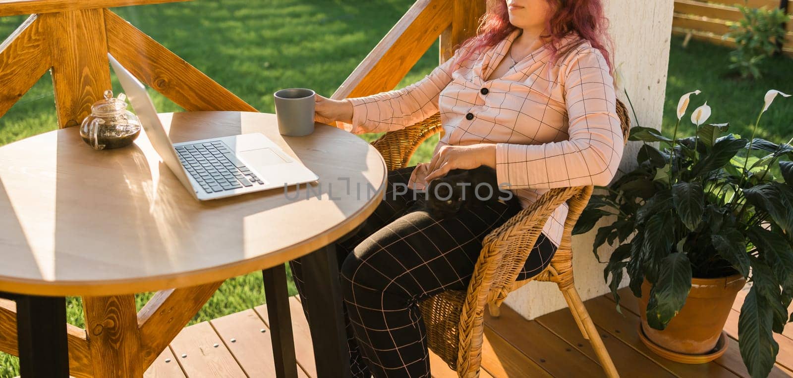 Woman working with laptop outdoor in summer terrace with black kitten. Girl and cat watching video on computer in the evening. Working from home, business, pet and lifestyle