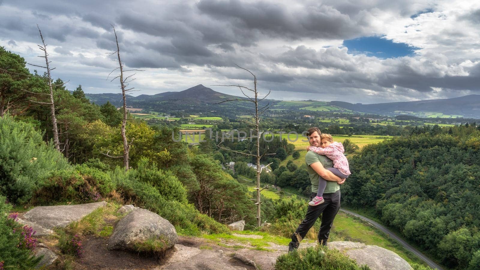 Father holding daughter in arms and standing on edge of cliff, with a scenic view on a valley and pine forest. Family hike, Wicklow Mountains, Ireland