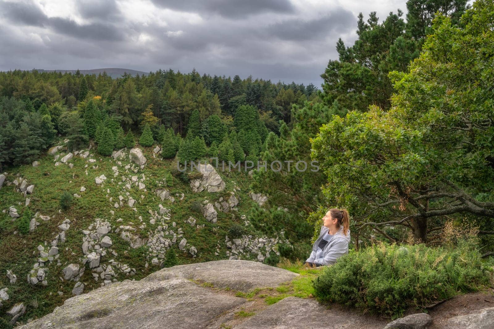 Adult woman leaning over a rock in Lead Mines, with a beautiful view on a valley and forest in background. Hiking in Wicklow Mountains, Ireland
