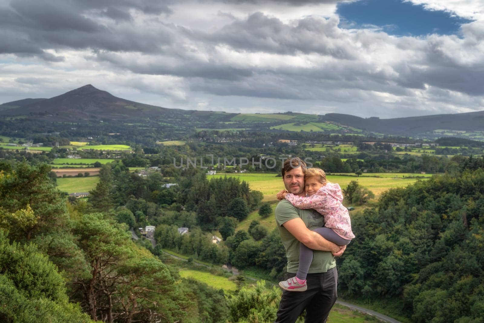 Father holding his daughter in arms with a scenic view on a valley, forest and mountain range. Family hike in Lead Mines, Wicklow Mountains, Ireland