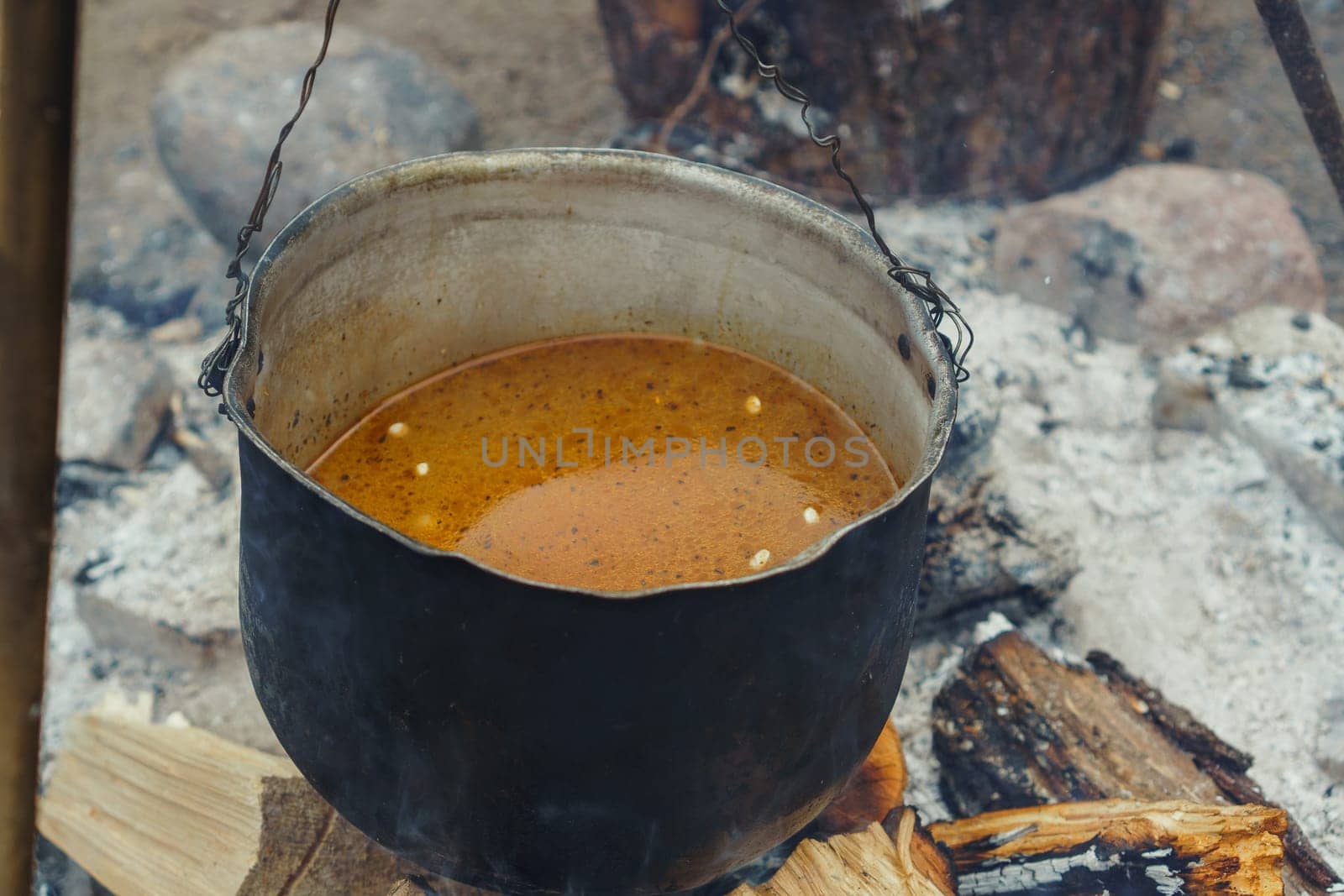 Metal vessel with spicy and mouthwatering soup hanging on tripod under firewood before cooking on fire in nature. Orange liquid meal for camping preparing in cauldron in village.
