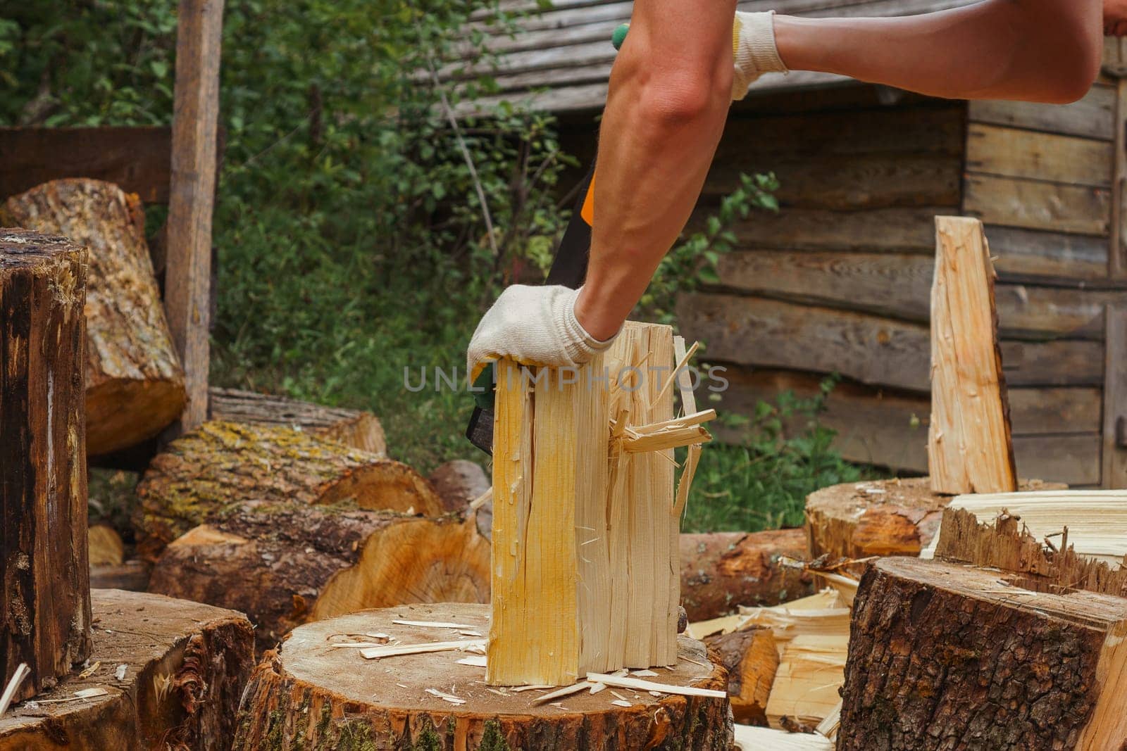 Unrecognizable adult man in gloves preparing firewood for campfire, holding log with hand and chopping wood using ax. Strong man cutting trees for camping bonfire, standing against village house.